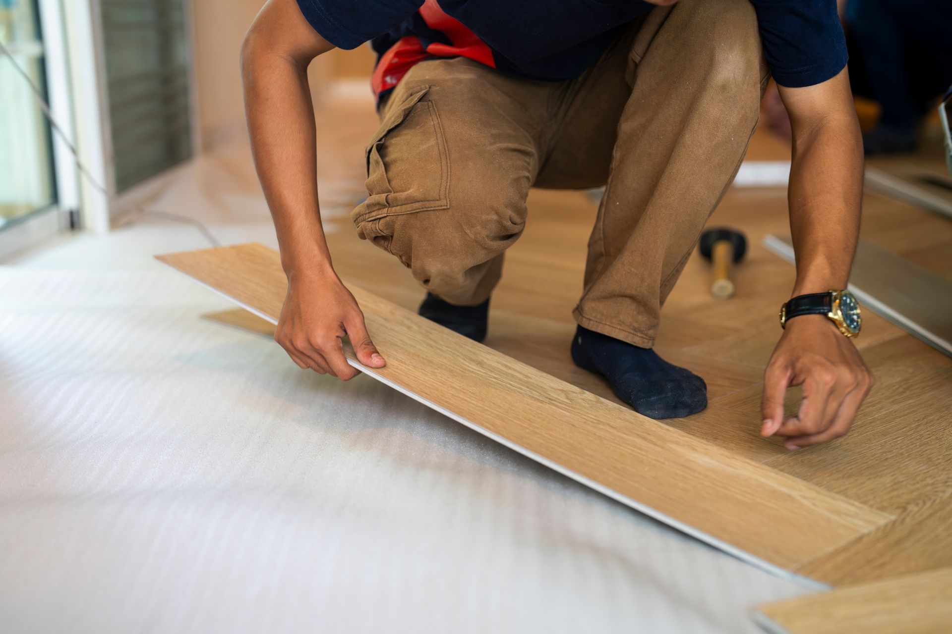 Person installing wood flooring indoors, kneeling on the floor, holding a plank, light brown and beige colors.