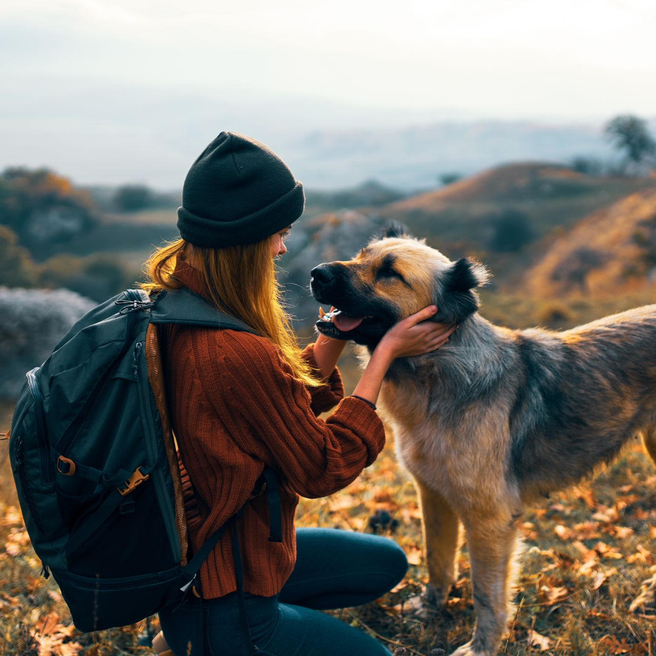 Woman petting a dog on a hillside. She wears a backpack and a knit cap. The dog is tan and smiling.