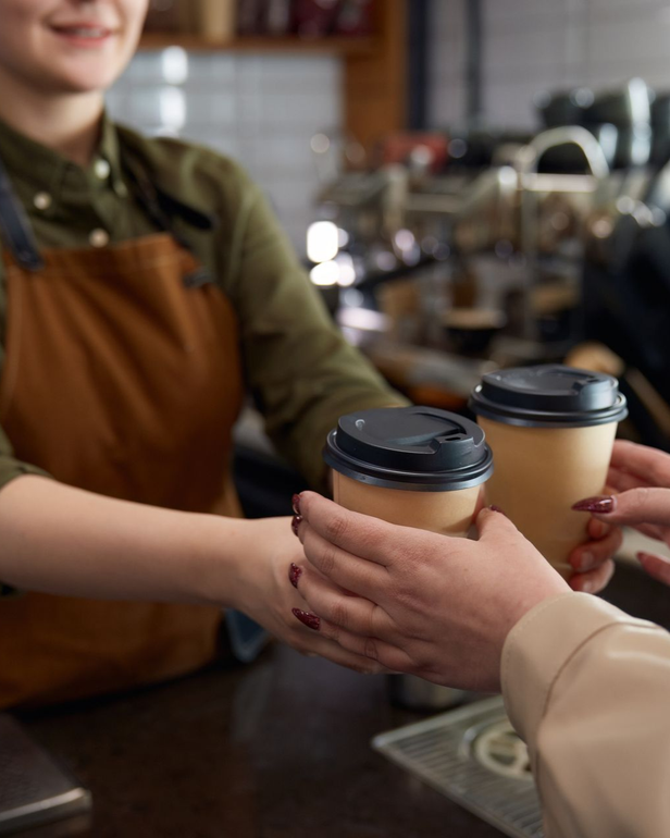 A barista in brown apron hands two coffees to a customer at a coffee shop.