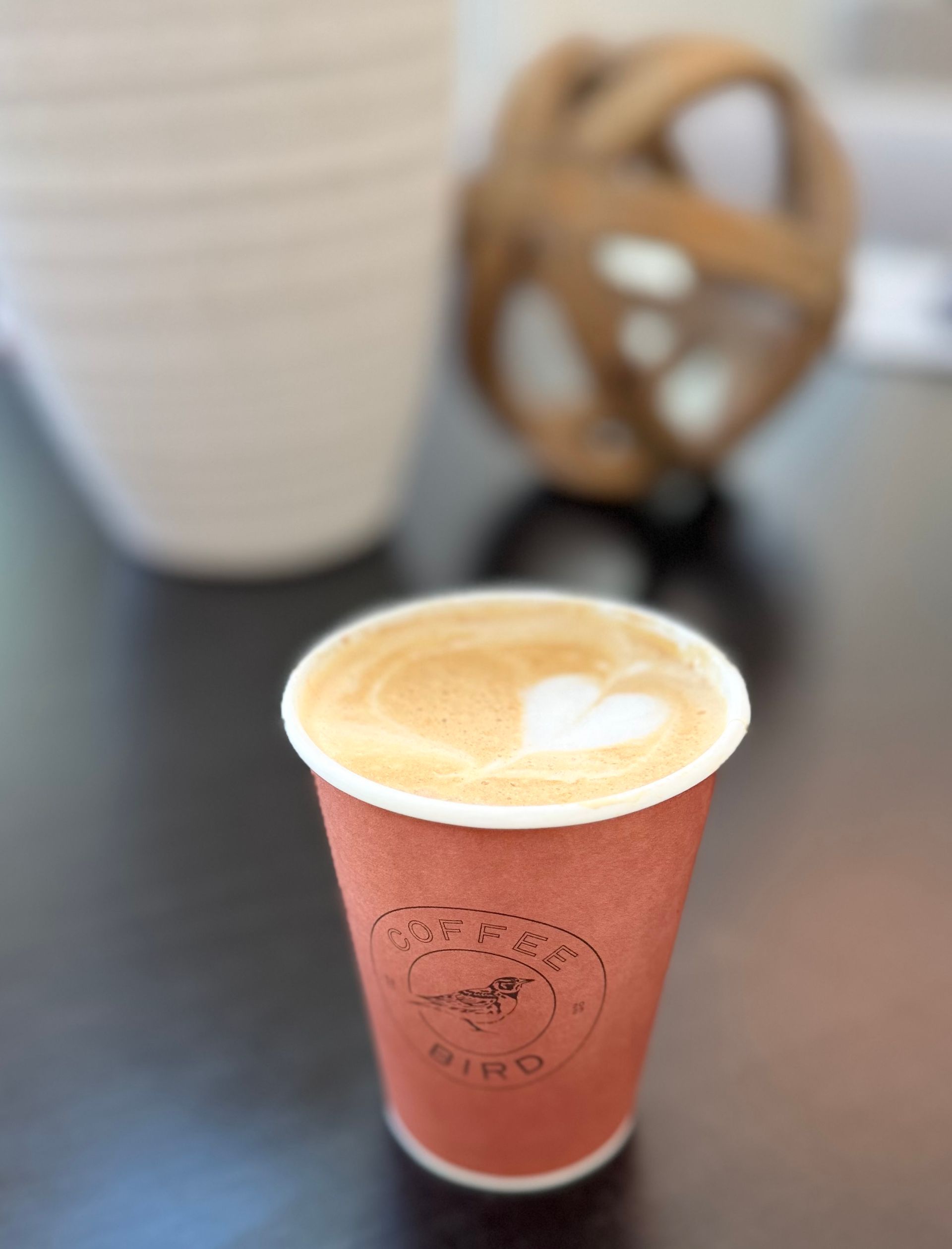 Coffee in a brown paper cup with latte art; set on a dark surface, with blurred decor in background.