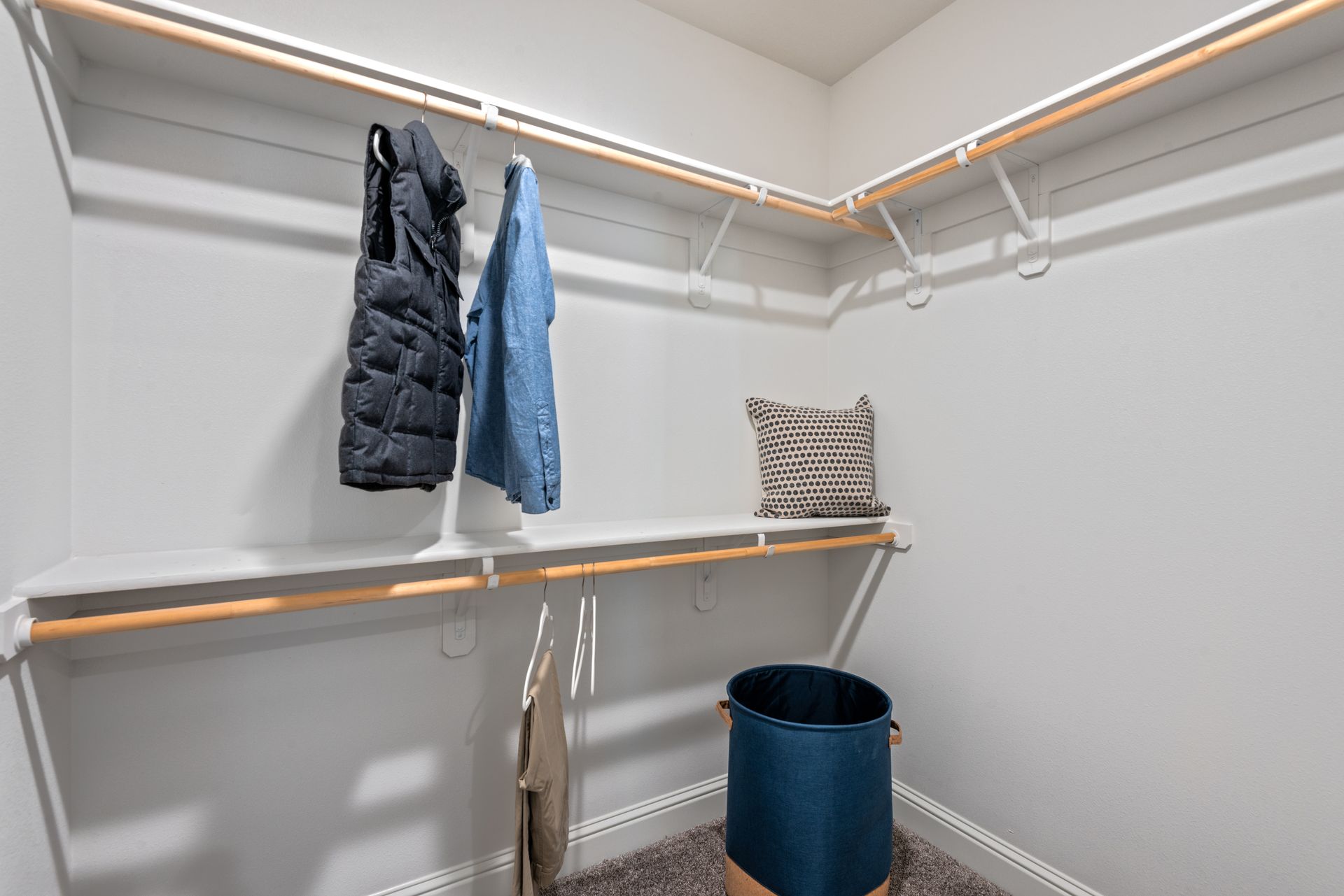 A walk-in closet with clothes hanging, a pillow, and a laundry basket. White shelves and rods.