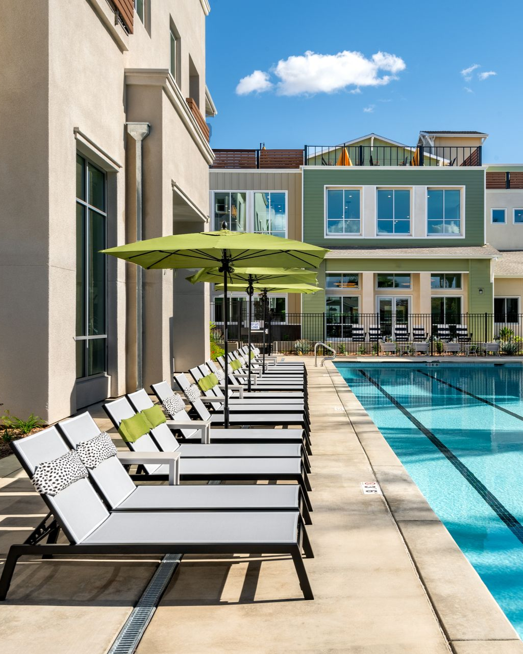 Poolside scene with lounge chairs, green umbrellas, and a building in the background. Blue sky and sunlight.