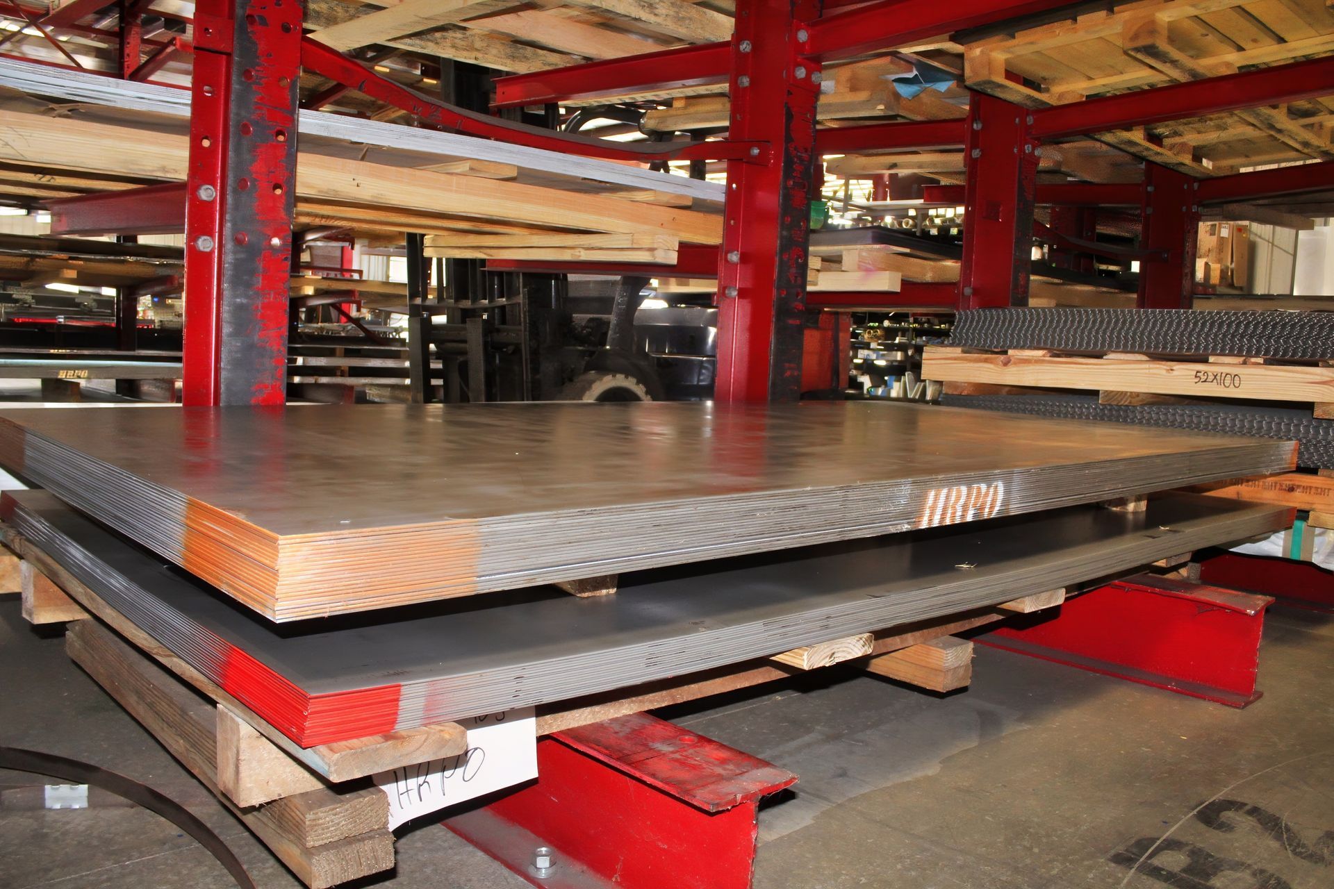 Metal sheets stacked on wooden pallets in a warehouse. Red and black shelving is visible.