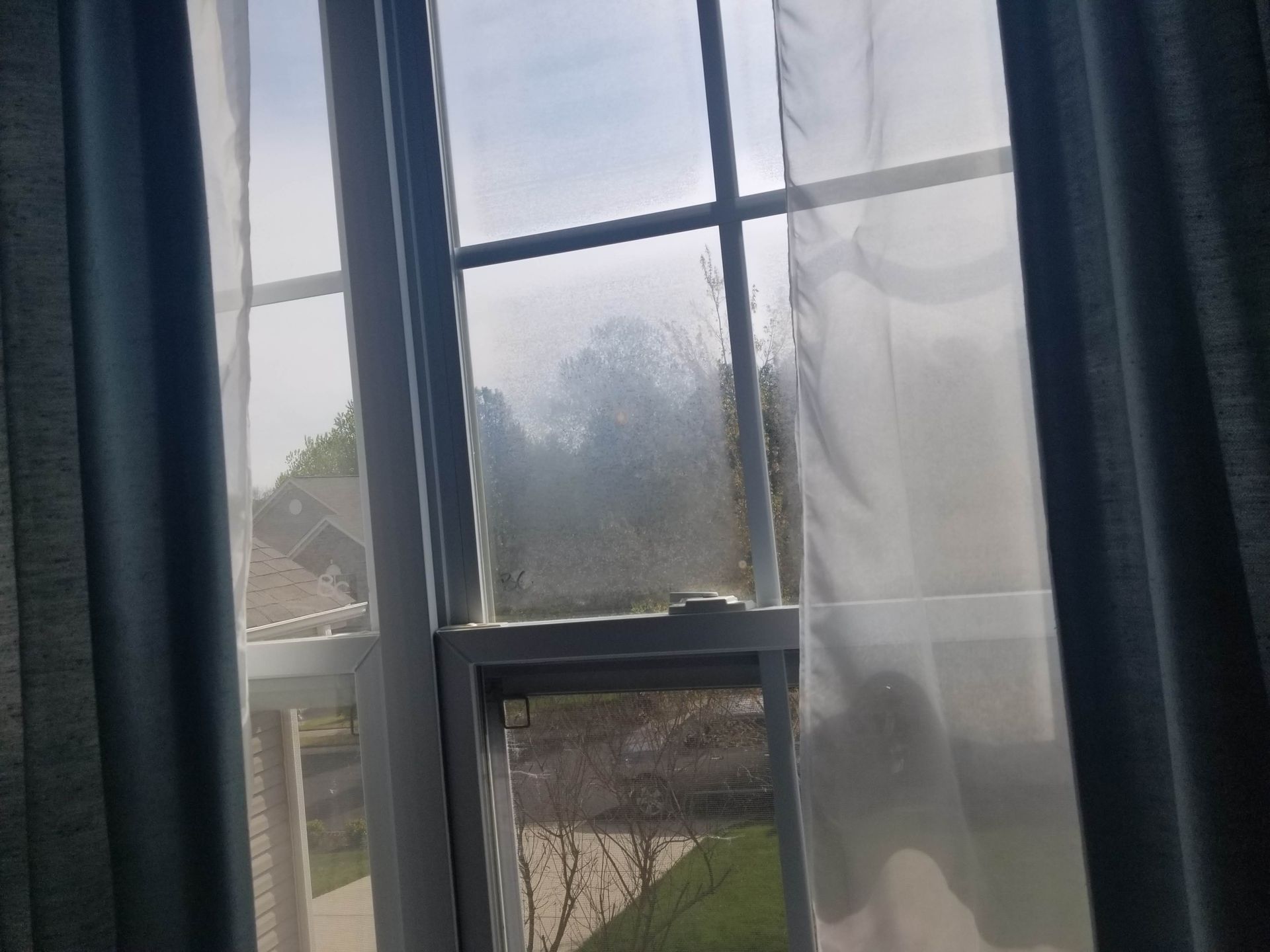 A window with sheer curtains is open and looking out to a yard.