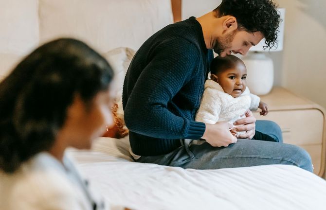 Man holding baby on bed; woman in foreground, neutral colors.