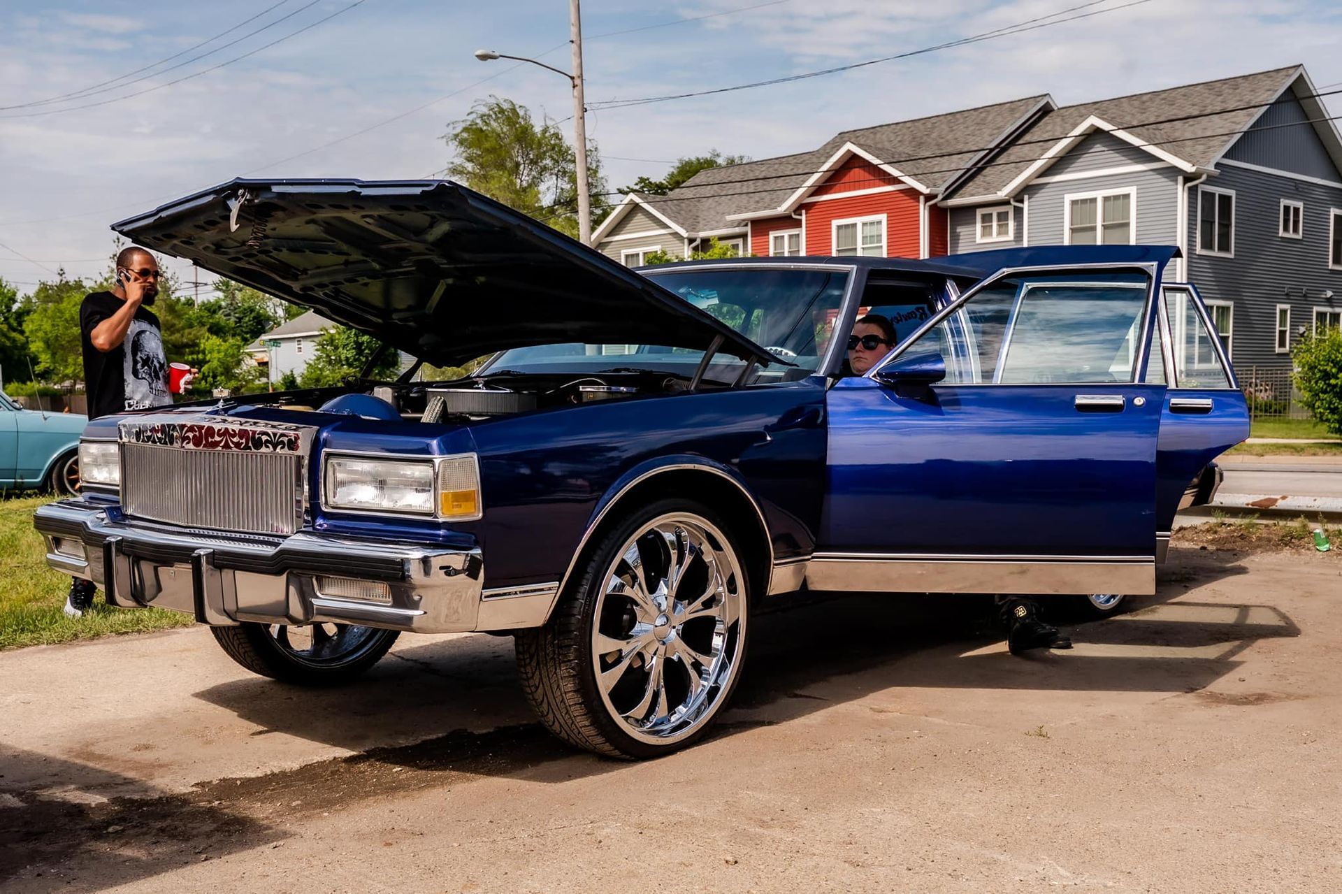 Blue classic car with chrome wheels, hood and door open, parked on a street.