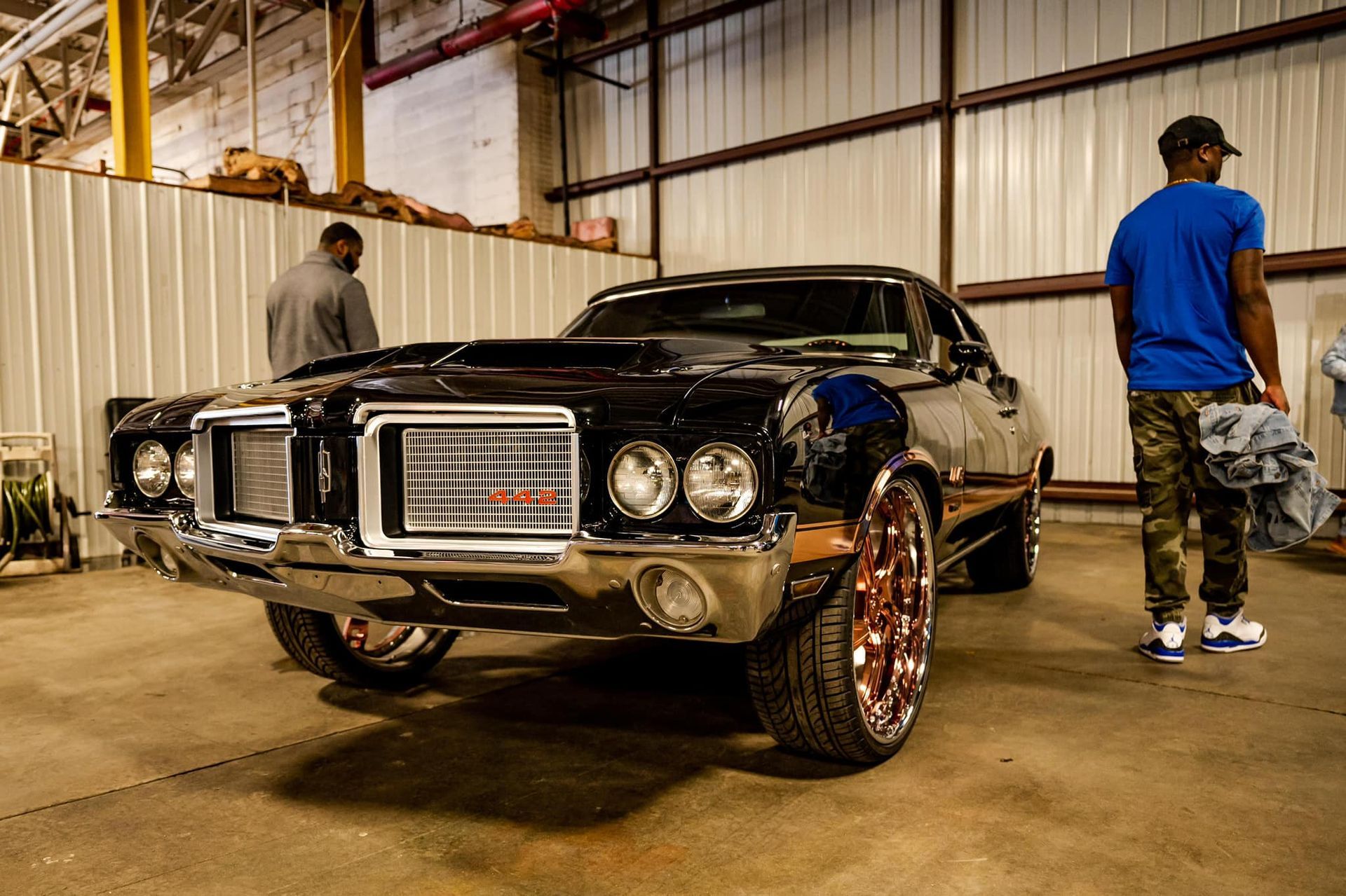 Black classic car with chrome accents inside a warehouse, people in background.