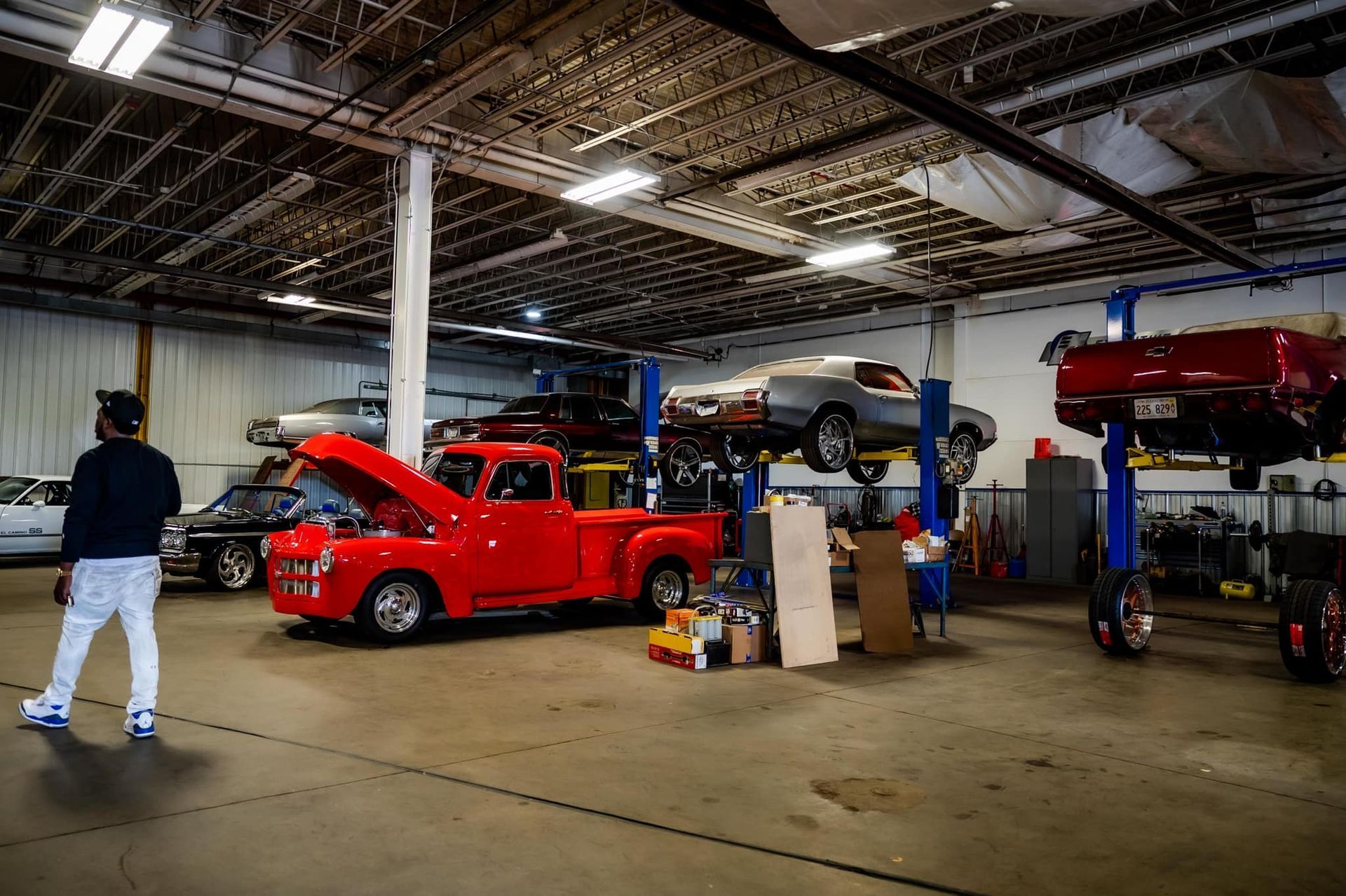 Interior of auto repair shop with classic cars on lifts, person looking at a red truck.