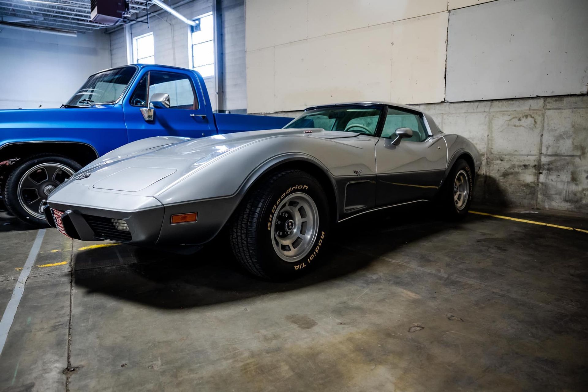 Silver Corvette parked next to a blue pickup truck in a garage.