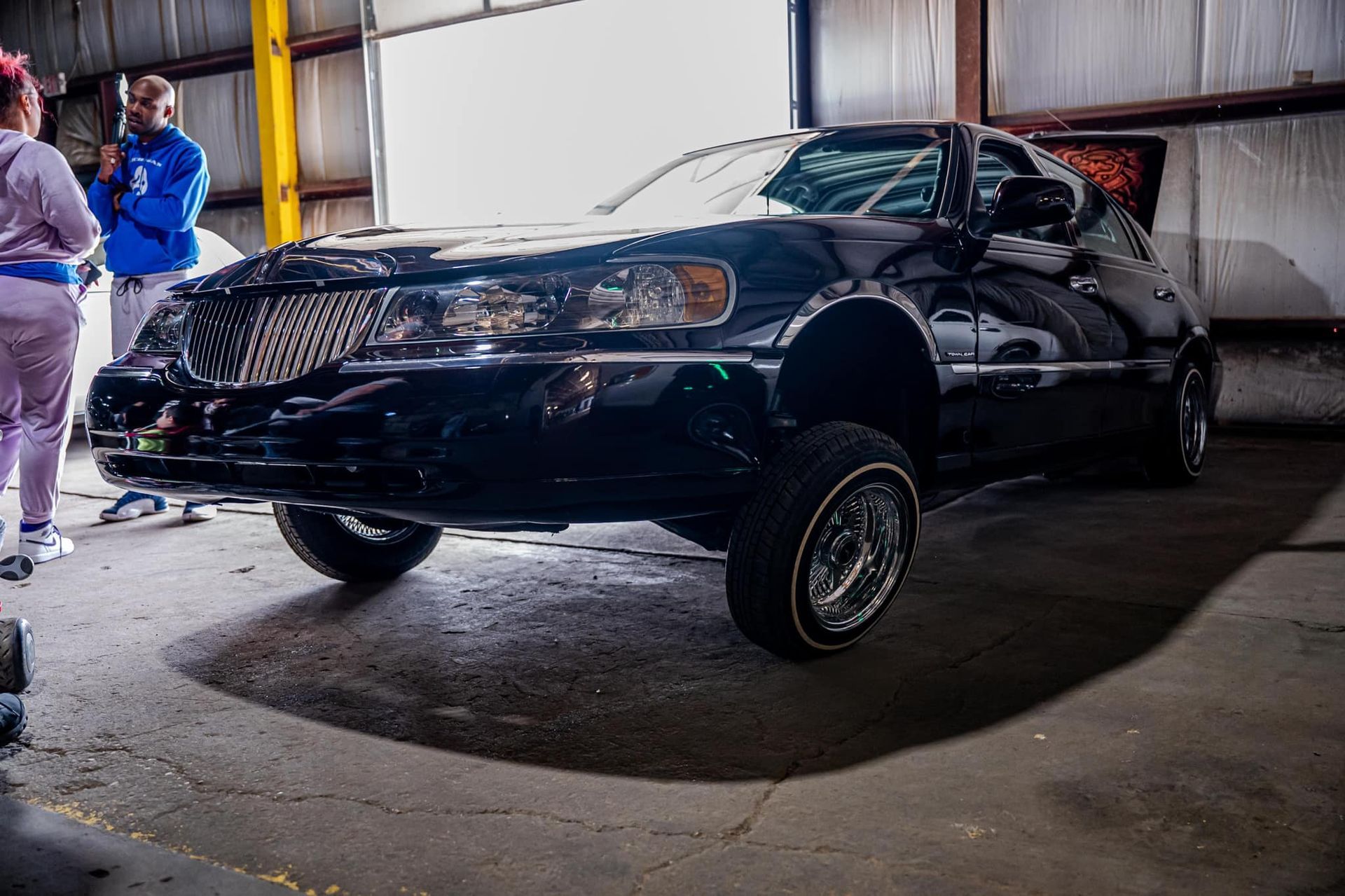 Black lowrider car, chrome details, raised suspension, indoors. People in background.