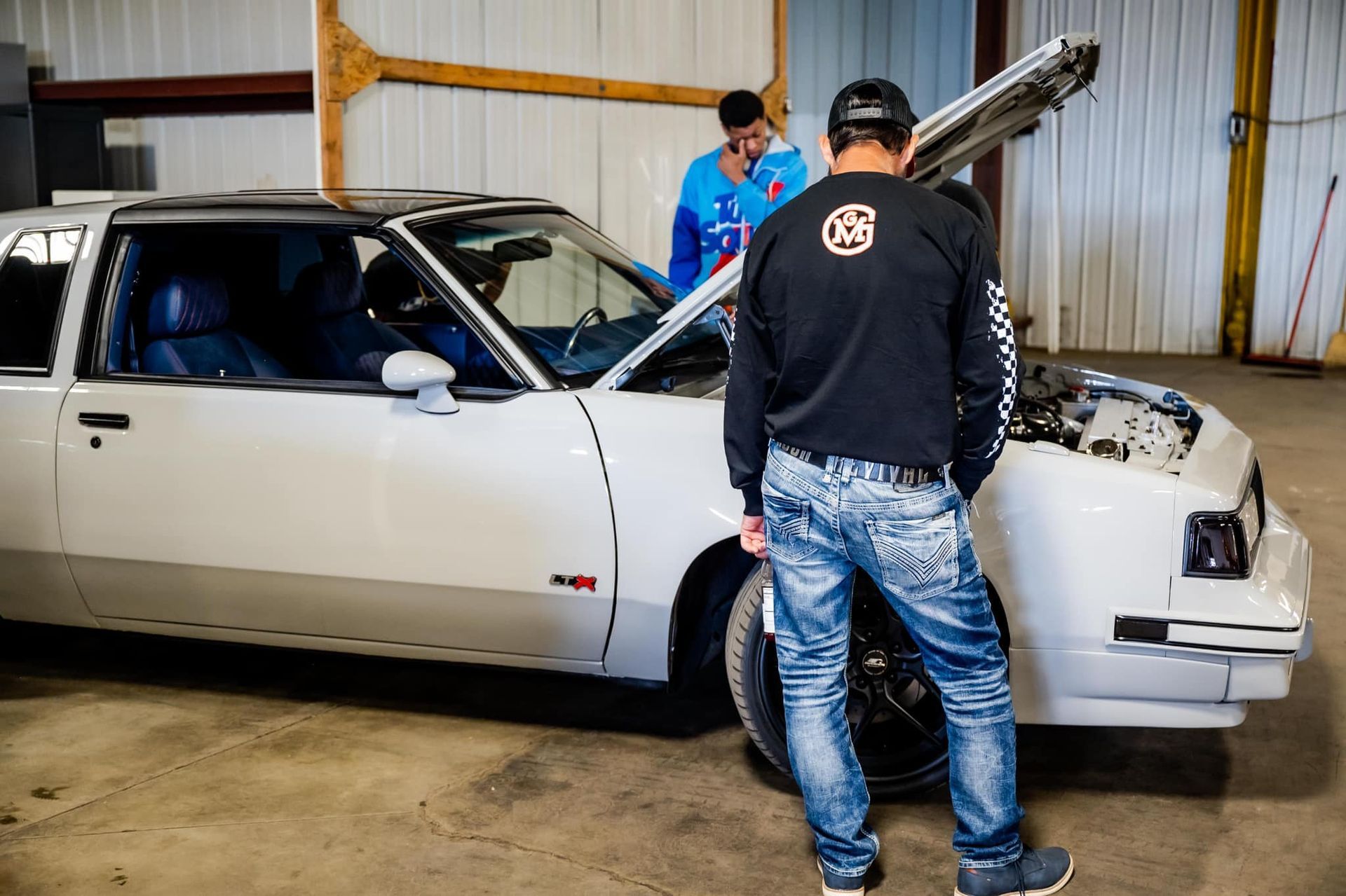 Two men inspecting a white car with an open hood inside a garage.