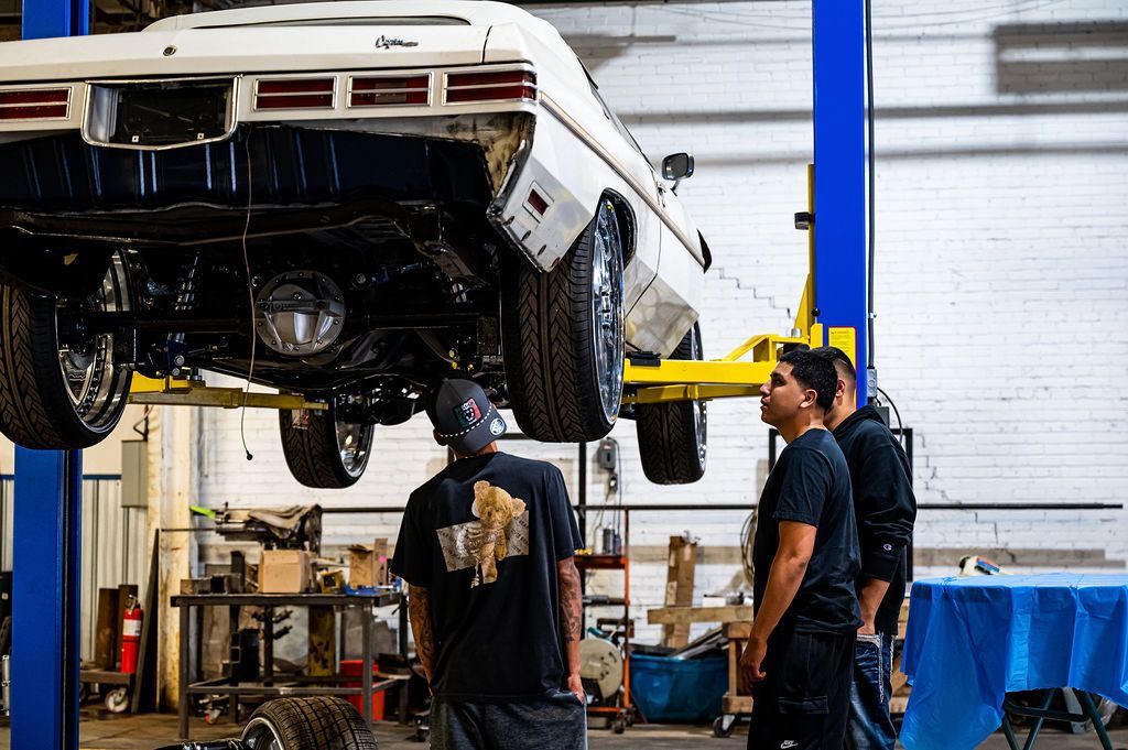 Car on a lift in a garage with three people looking underneath; one person is wearing a black shirt with a gold graphic.