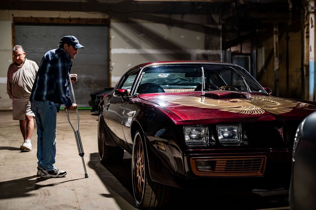 Man with crutches examines a red classic car in a dimly lit garage; another person watches.