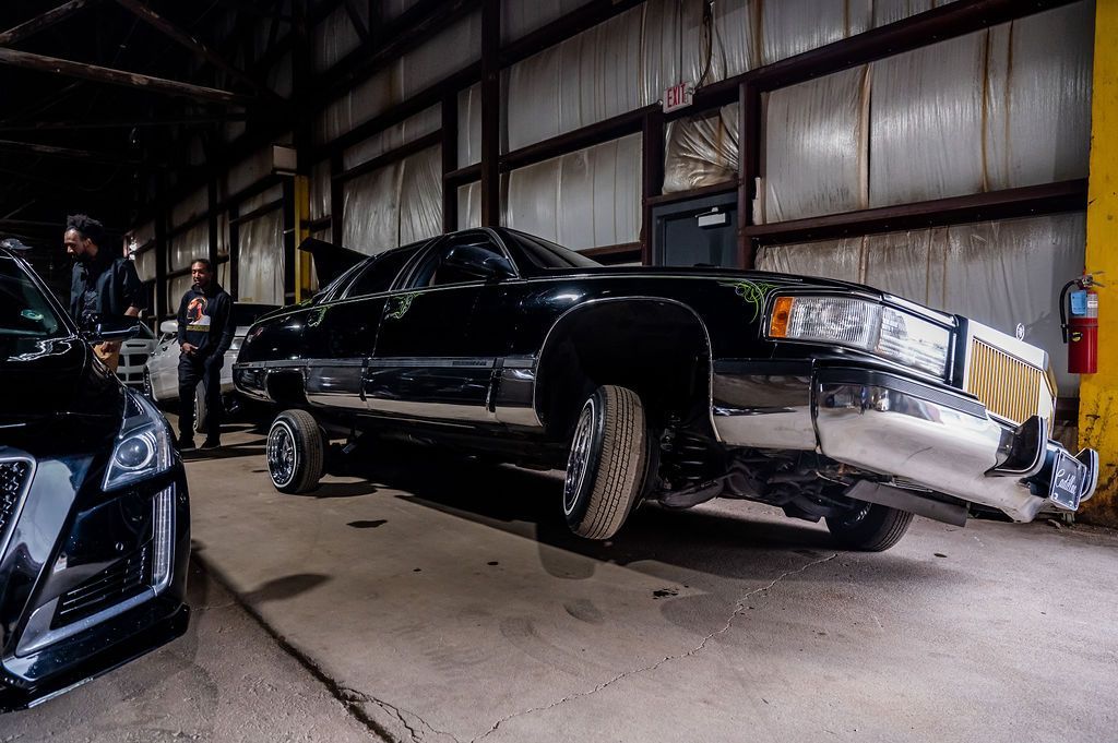 Black, modified limousine in a garage with people, chrome bumper and raised front.