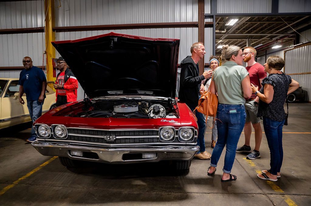 People gather around a red classic car with its hood open, likely at an indoor car show or event.