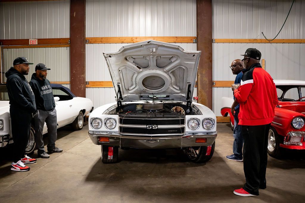 People examining a classic car with the hood open inside a garage. Other cars and garage interior are visible.