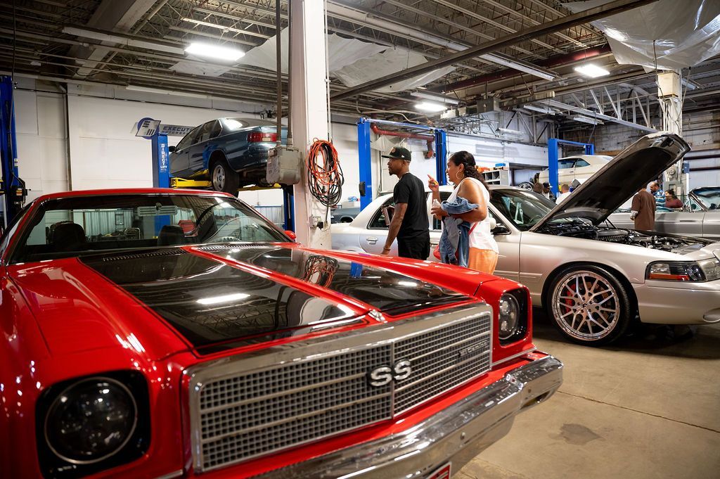 Classic cars in a garage. People inspecting a red car with black stripes, others near a car with an open hood.
