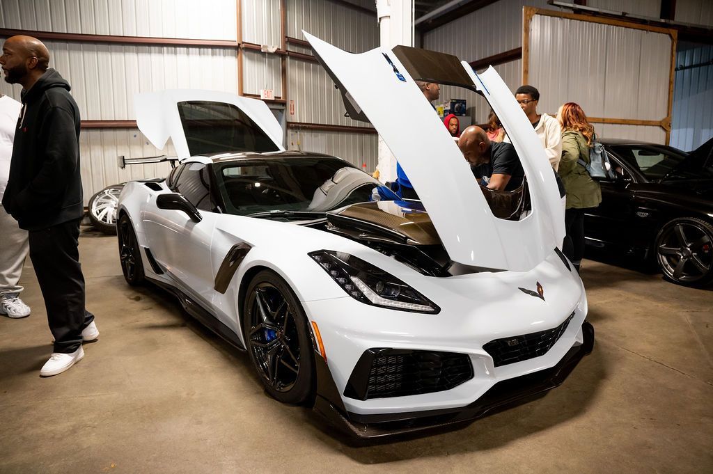 White sports car with open hood, displayed indoors with people looking on.