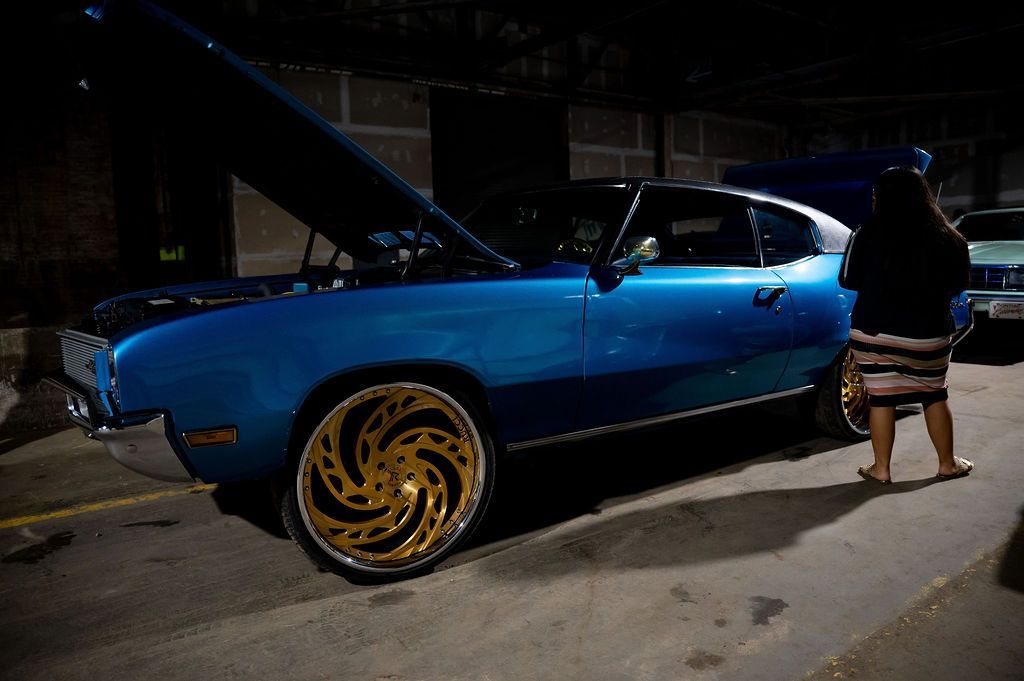 Blue classic car with open hood, gold rims, at night; person nearby.