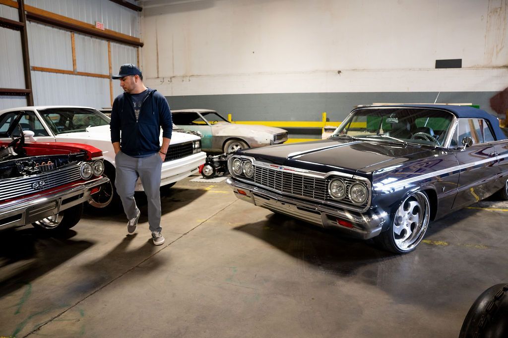 Man in garage with classic cars. One blue convertible, two others visible, inside a workshop.