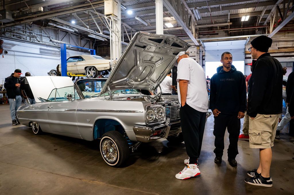 People admiring a silver lowrider car with the hood open inside a garage.