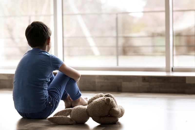 A young boy is sitting on the floor with a teddy bear and looking out a window.