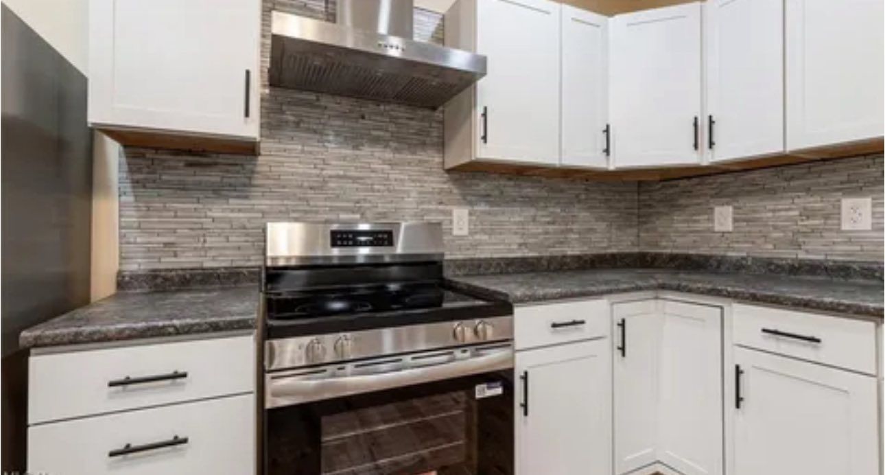A modern kitchen featuring white cabinets, stainless steel appliances, dark countertops, and a textured stone backsplash.