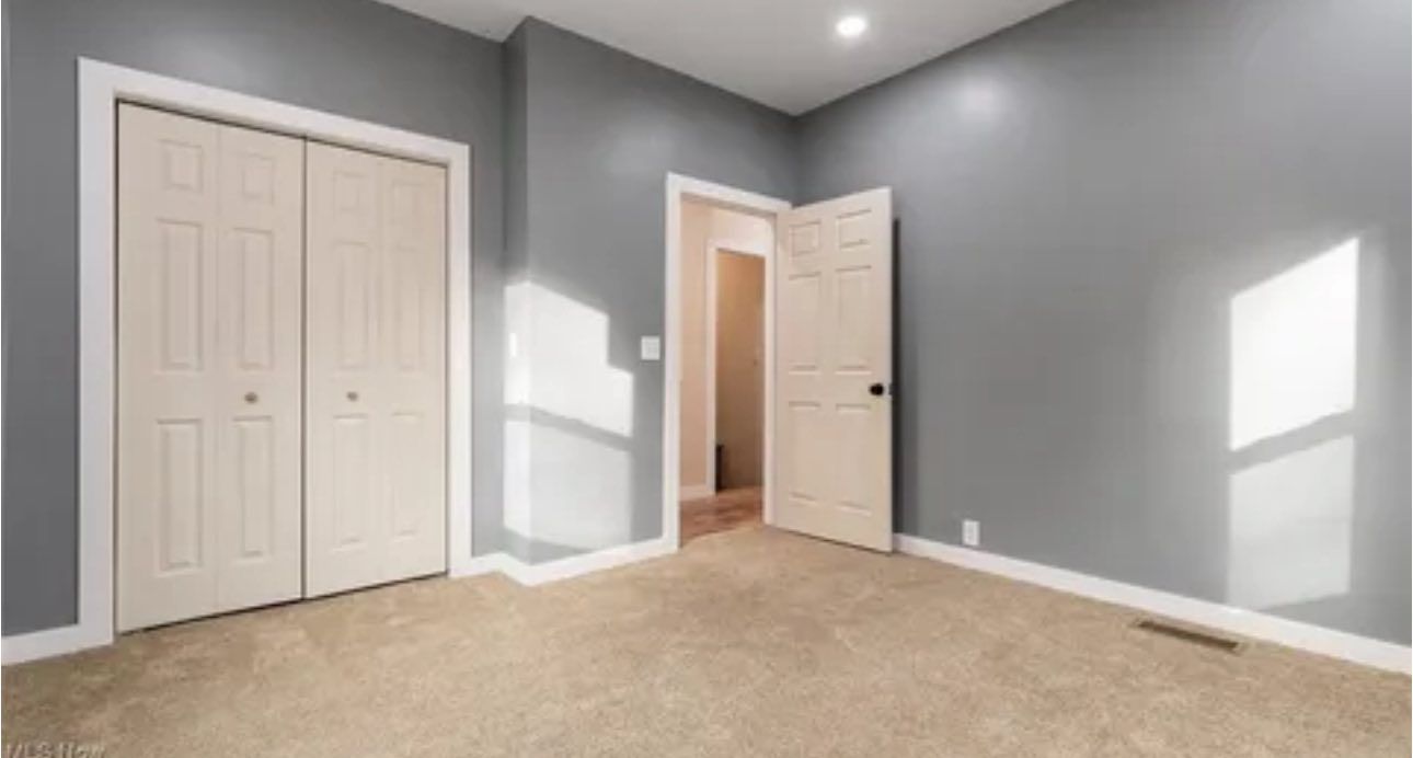 An empty room with gray walls, beige carpeting, a white sliding-door closet, and an open door to a hallway.