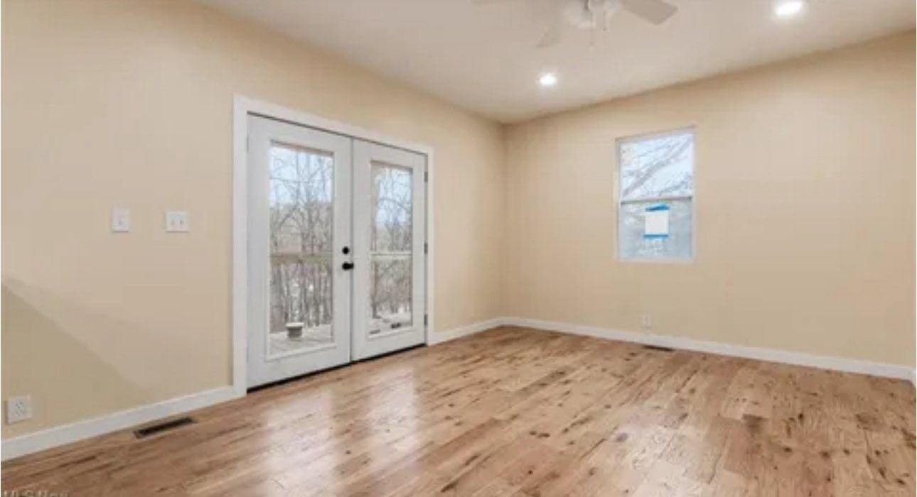 An empty room with light wood floors, beige walls, white trim, a glass French door, and a single window.