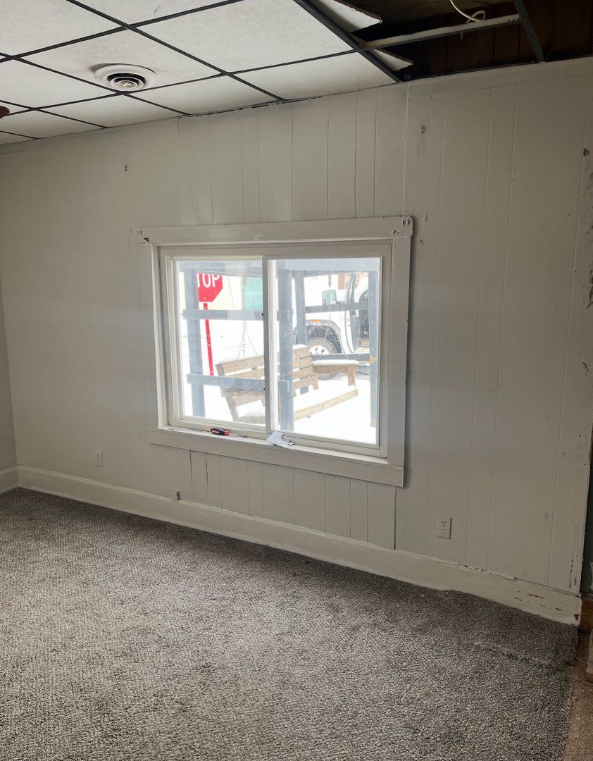 A room with white paneled walls, a single window looking outside, and grey carpet under a dropped ceiling grid.