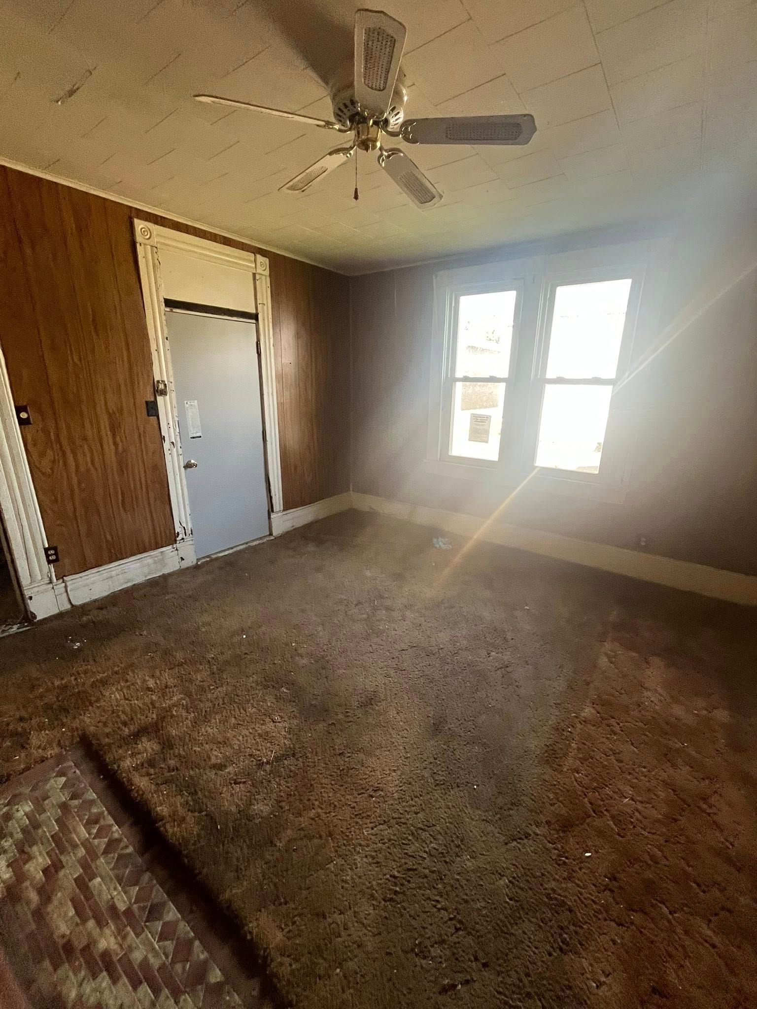 A dimly lit, unfurnished room with wood paneling, a ceiling fan, a grey door, and two windows bright with natural light.