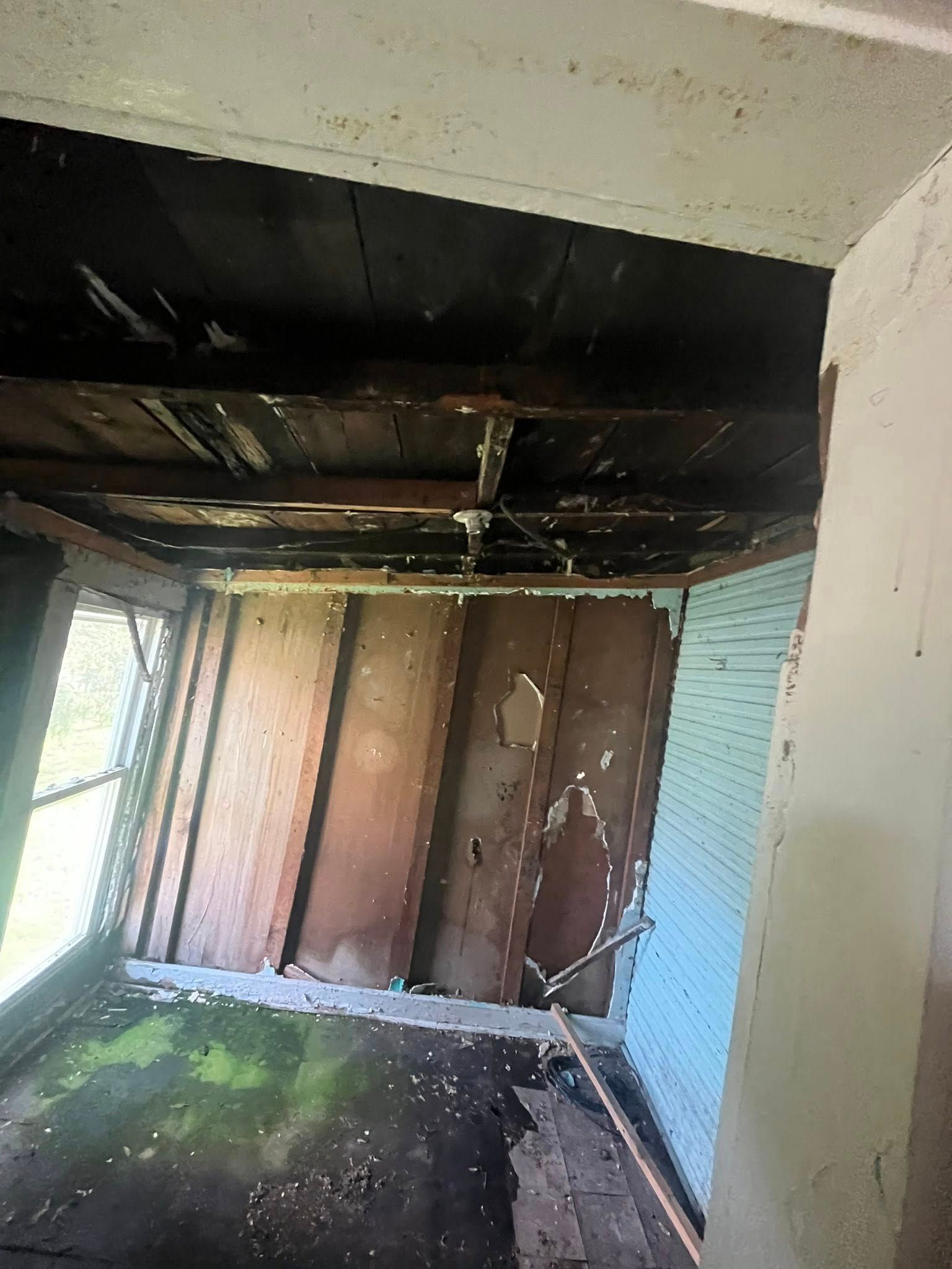 A dark, dilapidated porch with a charred ceiling, damaged wood walls, and a worn floor, viewed from an entryway.