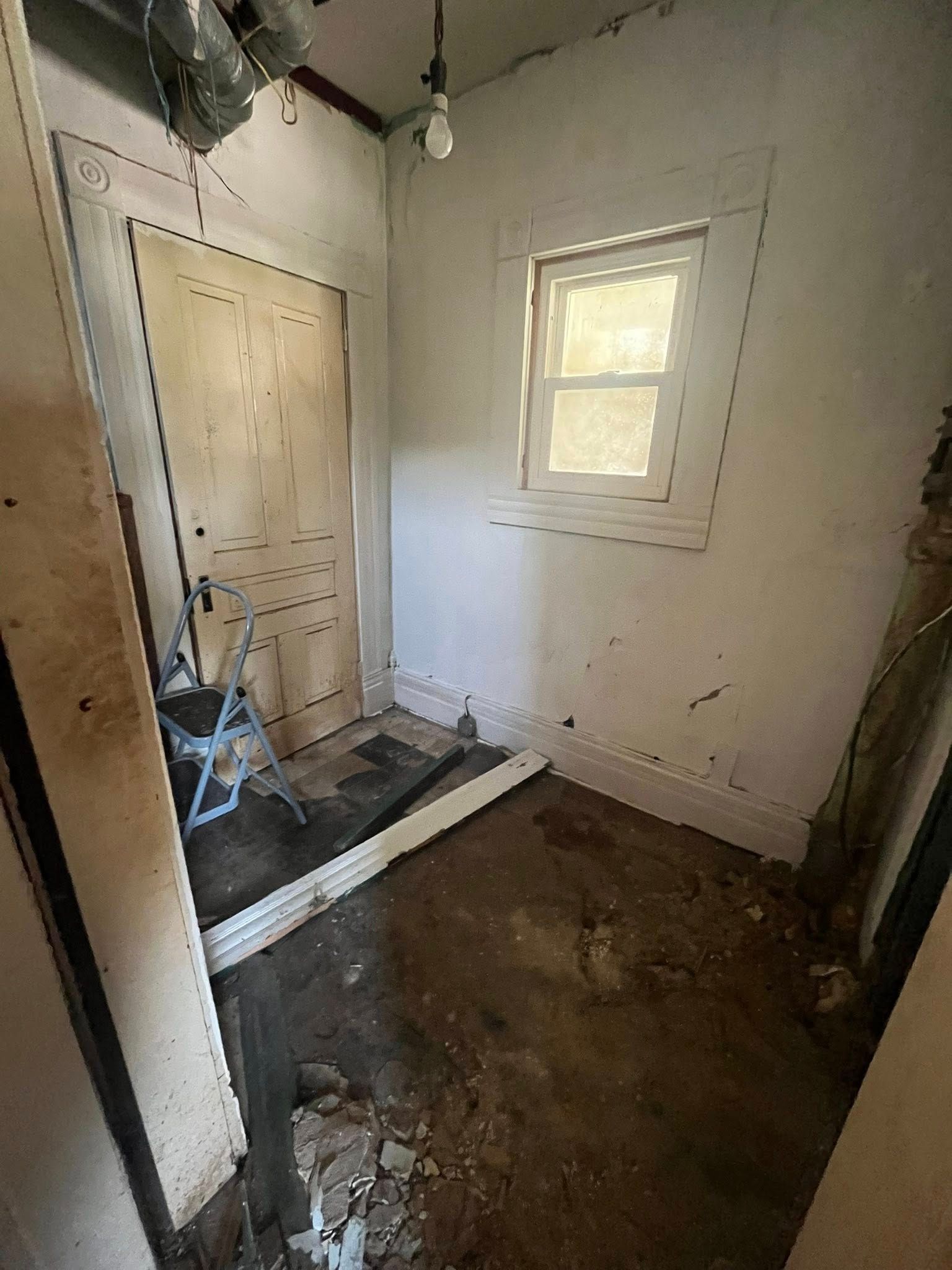 A dimly lit, unfinished room with a wooden door, a single window, exposed wires, and debris scattered on the floor.