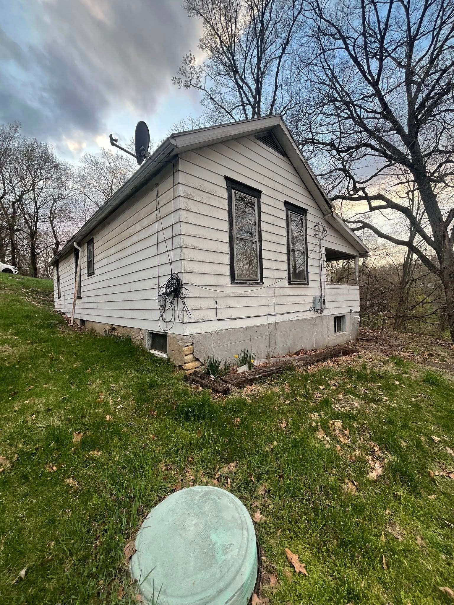 A small, white, weathered cottage with dark-trimmed windows sits on a grassy slope beneath bare trees at sunset.