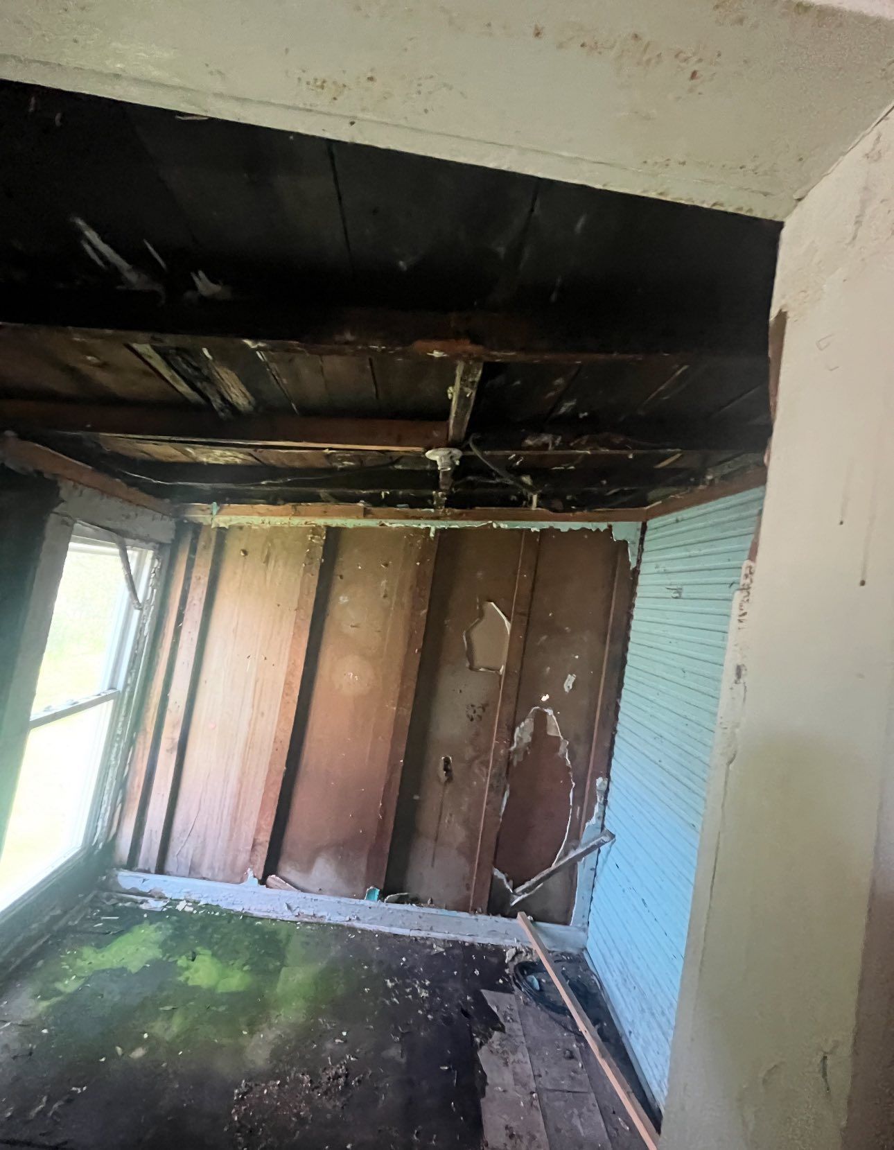 A dilapidated, dark room with exposed wooden ceiling beams, unfinished walls, and a moss-covered floor near a window.