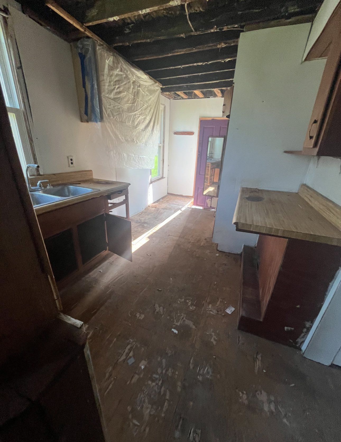 A dilapidated kitchen with exposed ceiling joists, wooden cabinets, a sink, and a doorway leading to another room.