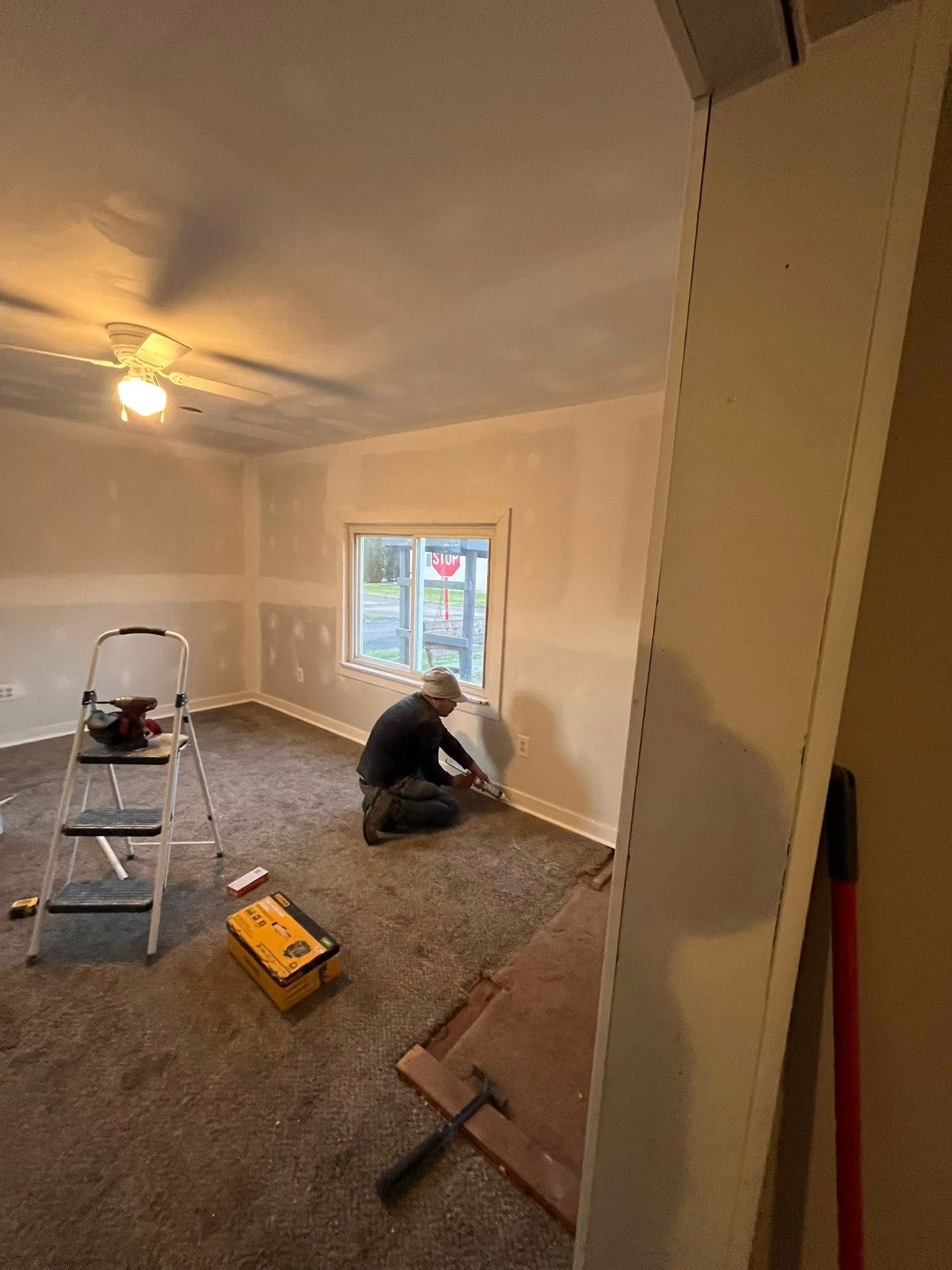 A person kneels on a carpeted floor in a room under renovation, working on baseboards beneath a window.