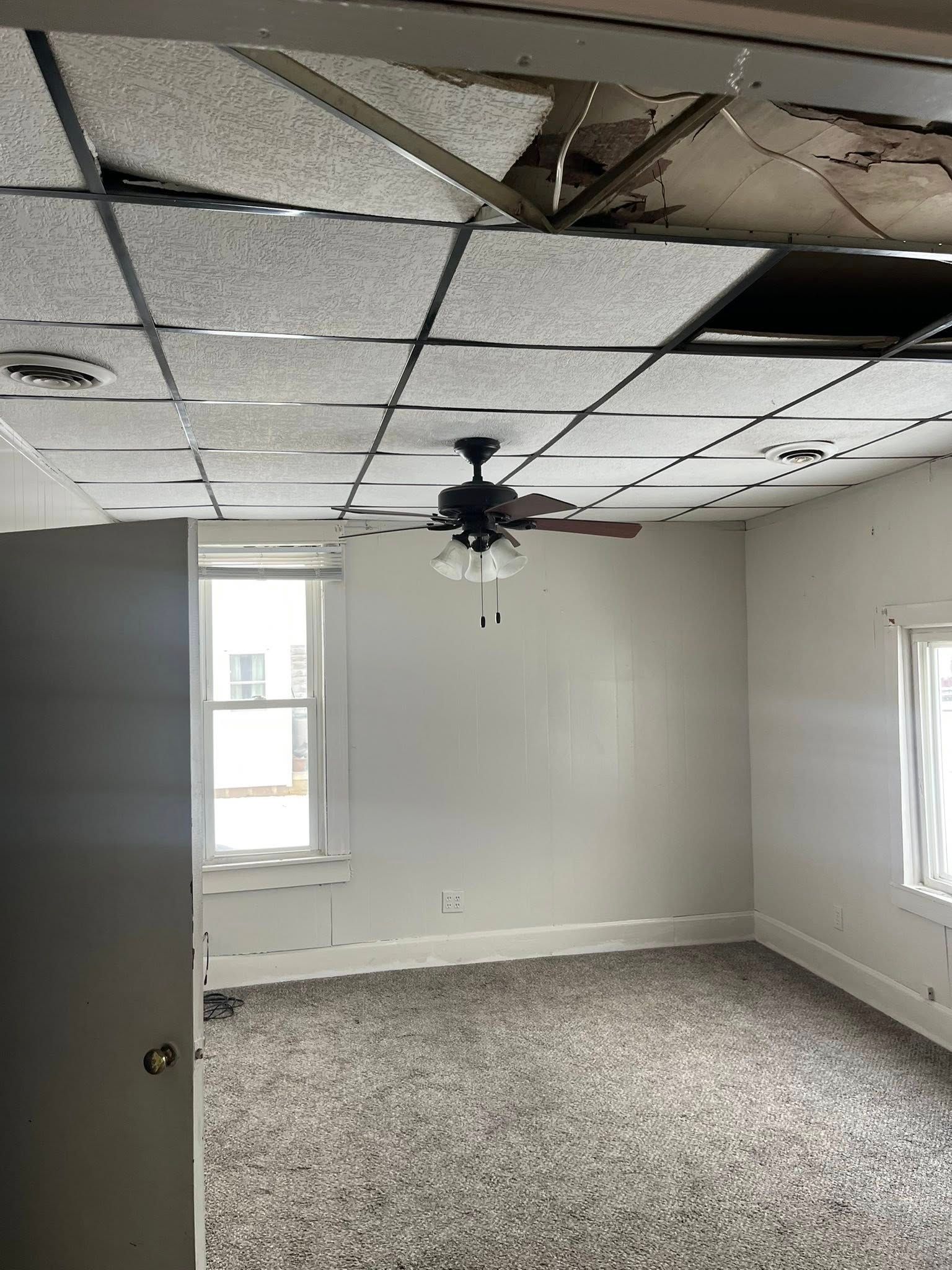 A sparsely furnished room with a gray patterned carpet, white walls, and a damaged suspended ceiling with a ceiling fan.