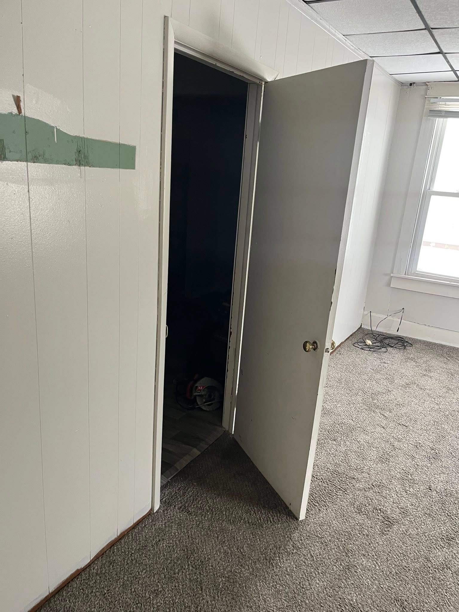 A white door stands slightly open, revealing a dark closet, in a room with white paneled walls and speckled gray carpet.