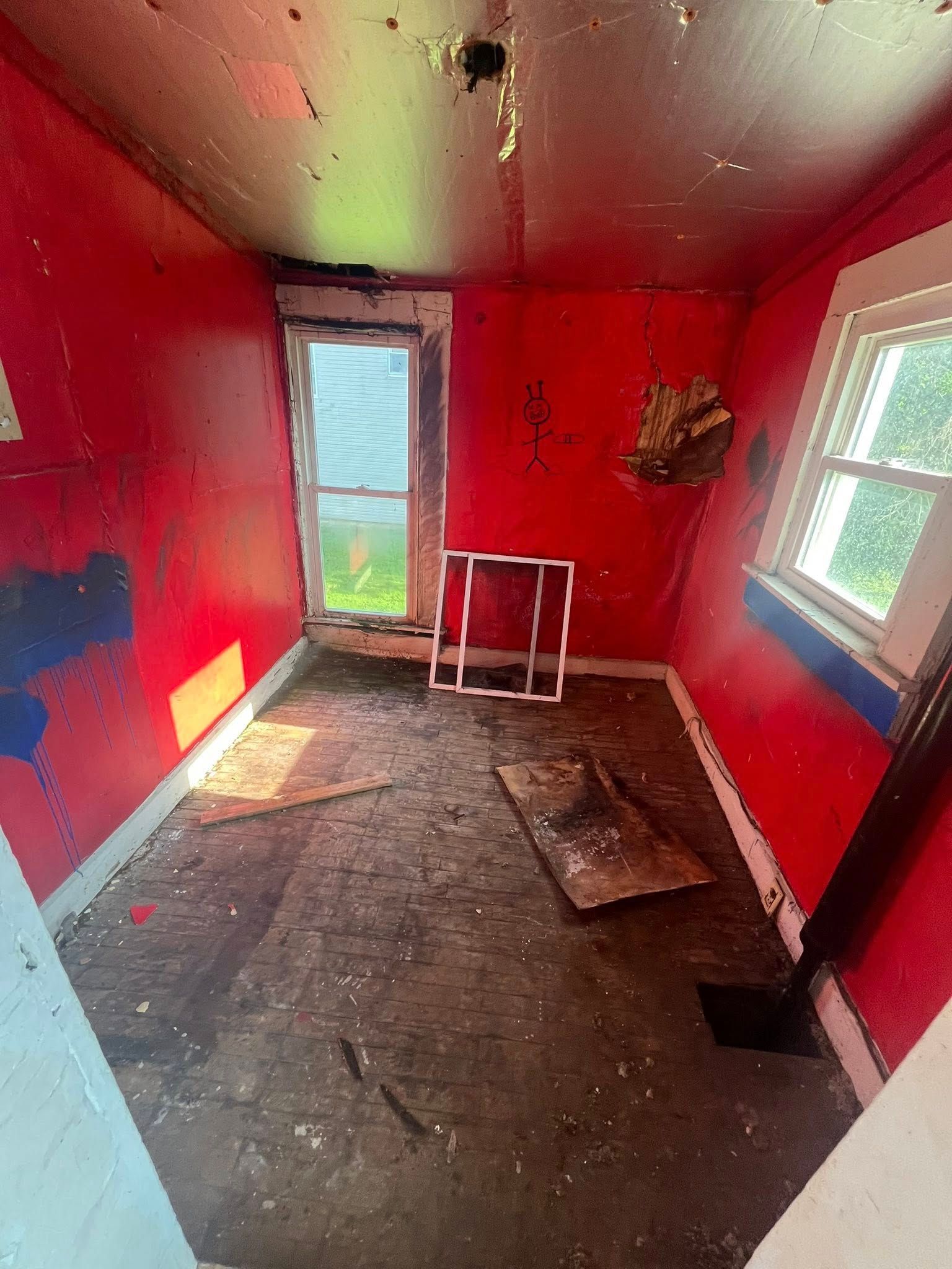 A dilapidated small room with bright red walls, a window on each side, exposed ceiling, and dark, debris-covered flooring.