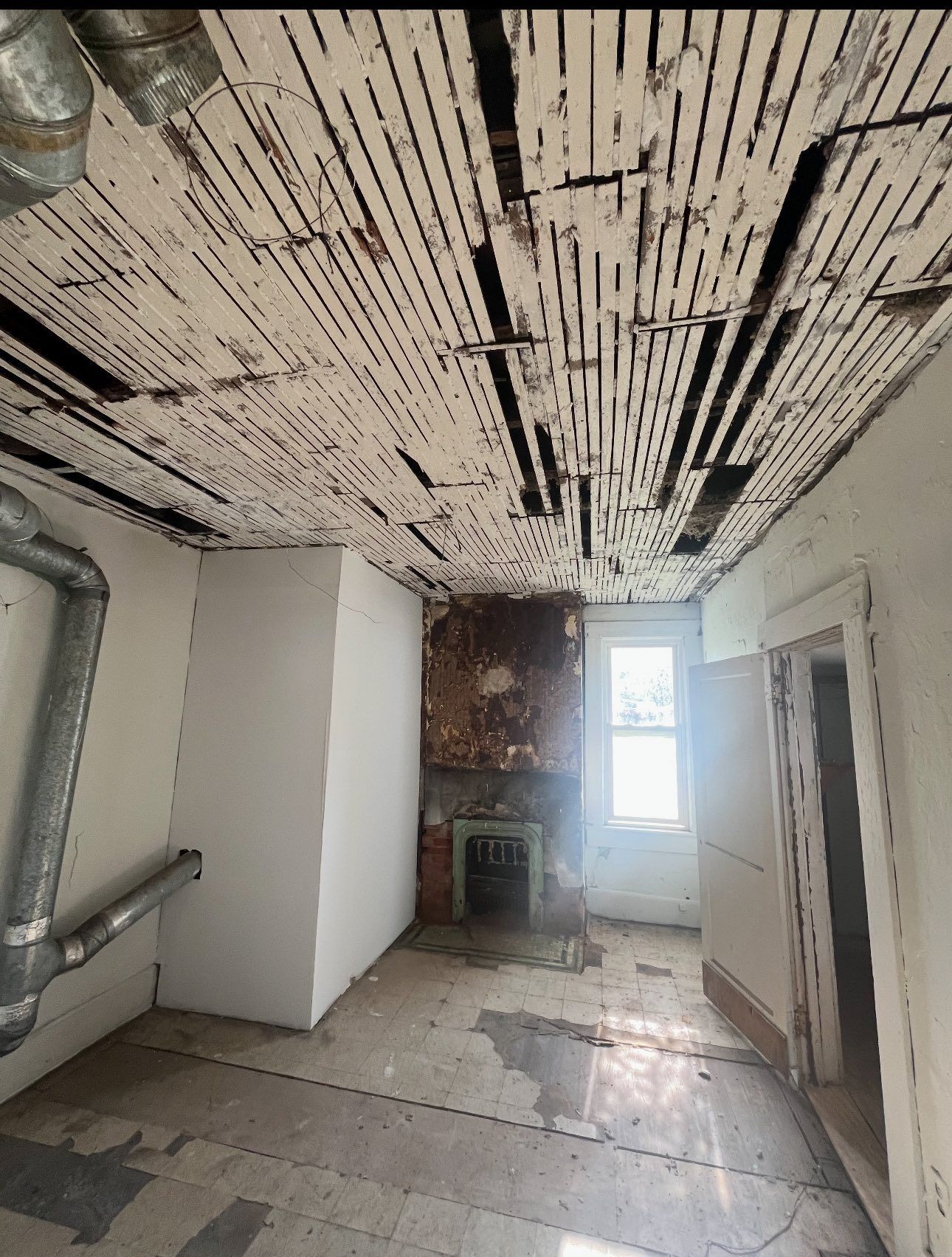 A dilapidated room with exposed wooden ceiling lath, bare walls, ductwork, and a fireplace at the far end by a window.