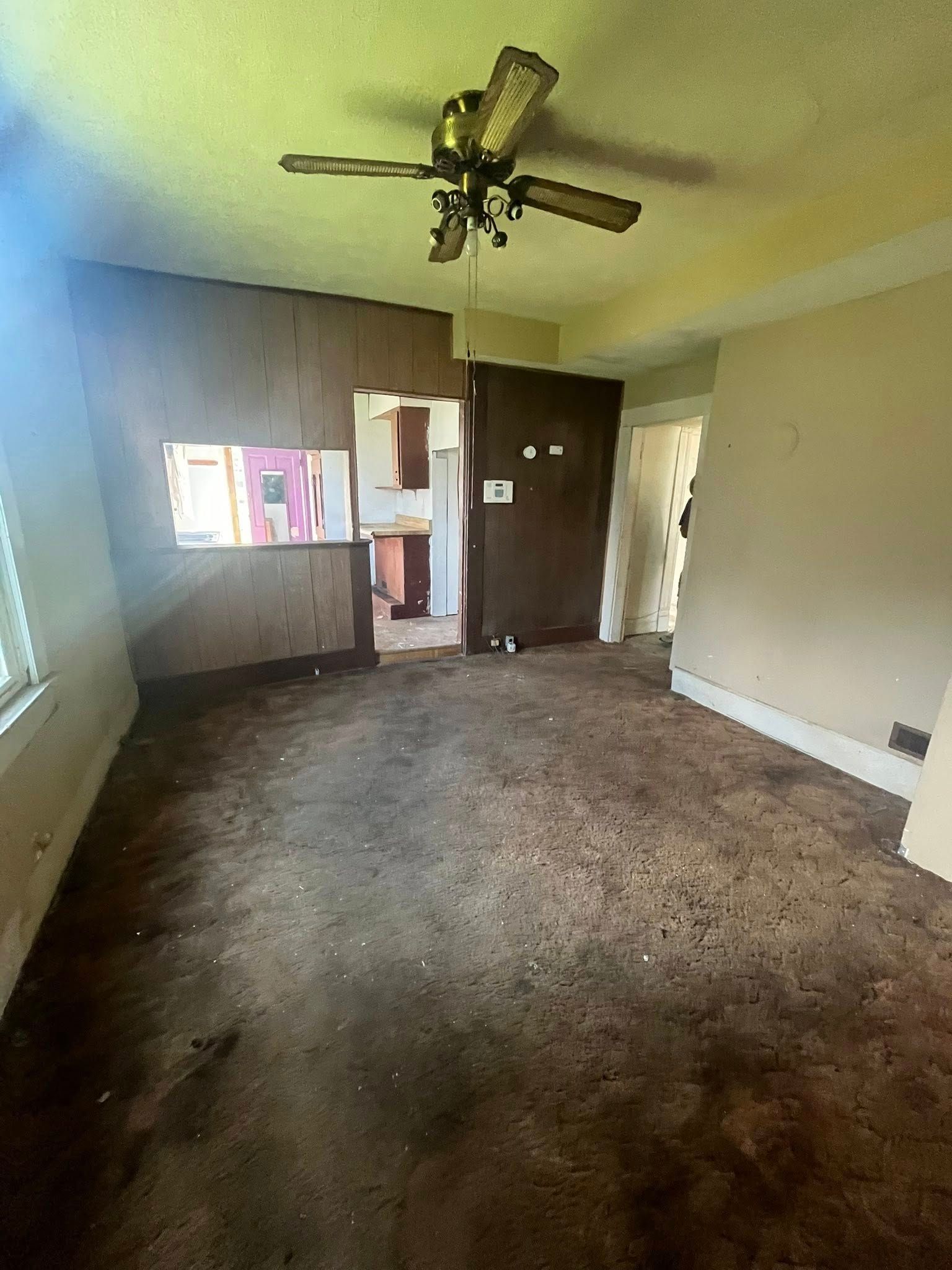 A dimly lit, unfurnished room with dark, stained carpeting, beige walls, a ceiling fan, and a doorway to a kitchen.