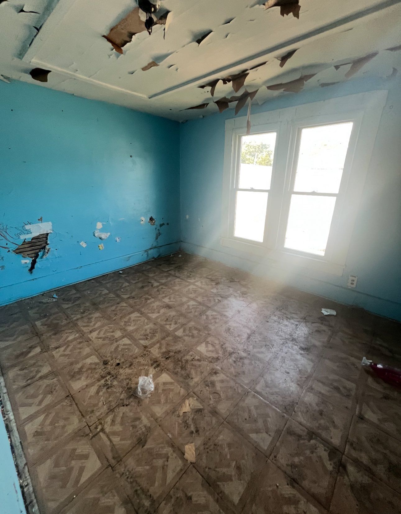A vacant room with bright blue walls in disrepair, peeling ceiling paint, and patterned tile flooring lit by a window.