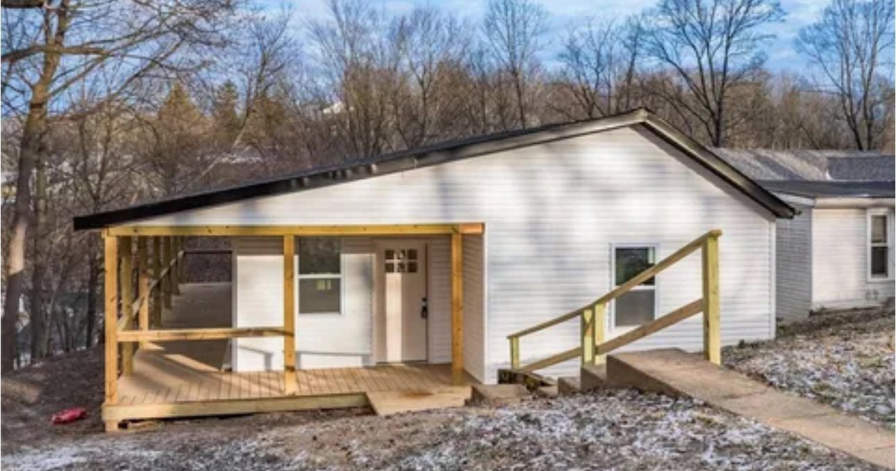 A white, single-story house with a wooden porch and deck, nestled in a wooded, hilly landscape on a sunny day.