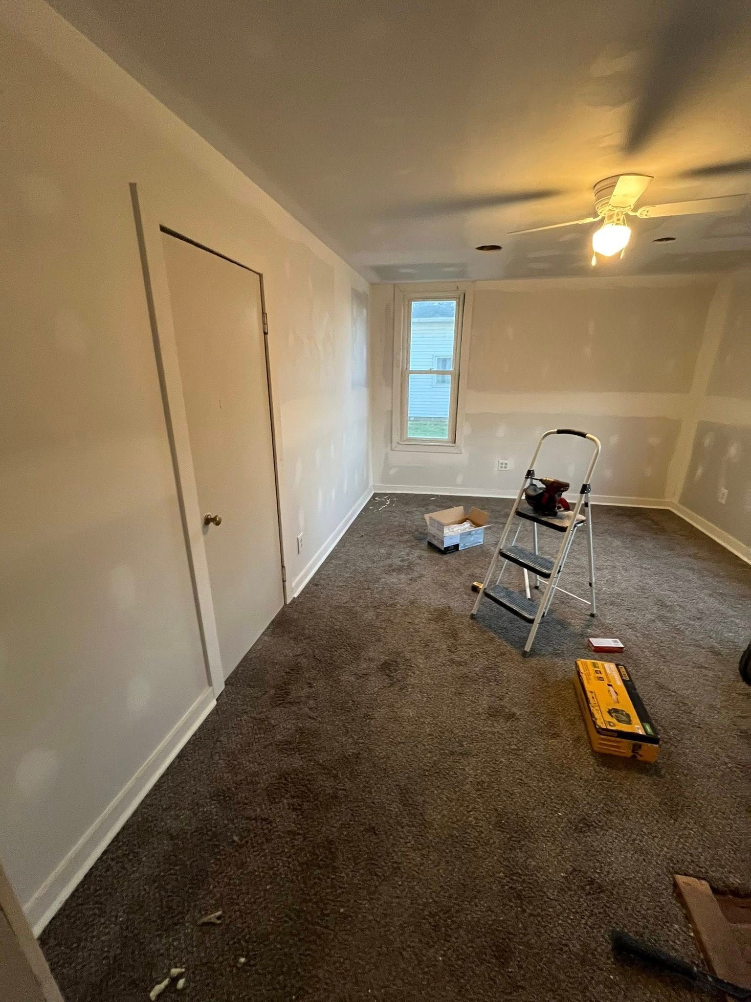 A bedroom under renovation with beige walls, a closed door, gray carpet, a metal ladder, and a yellow toolbox.