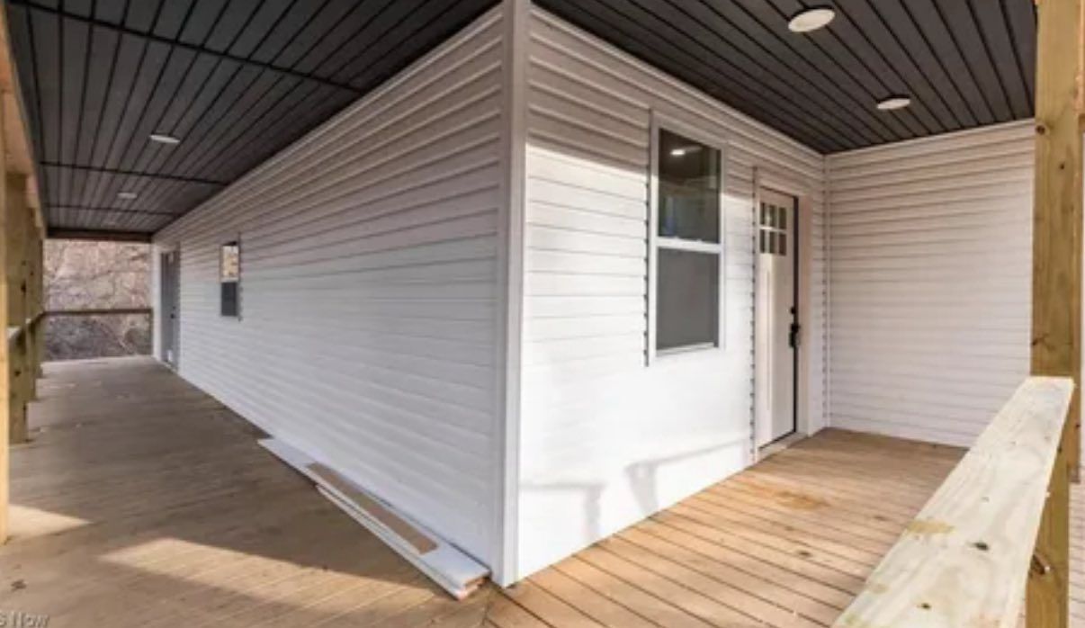 A wide exterior view of a white, vinyl-sided cabin with a wooden deck and a covered porch area under a dark ceiling.