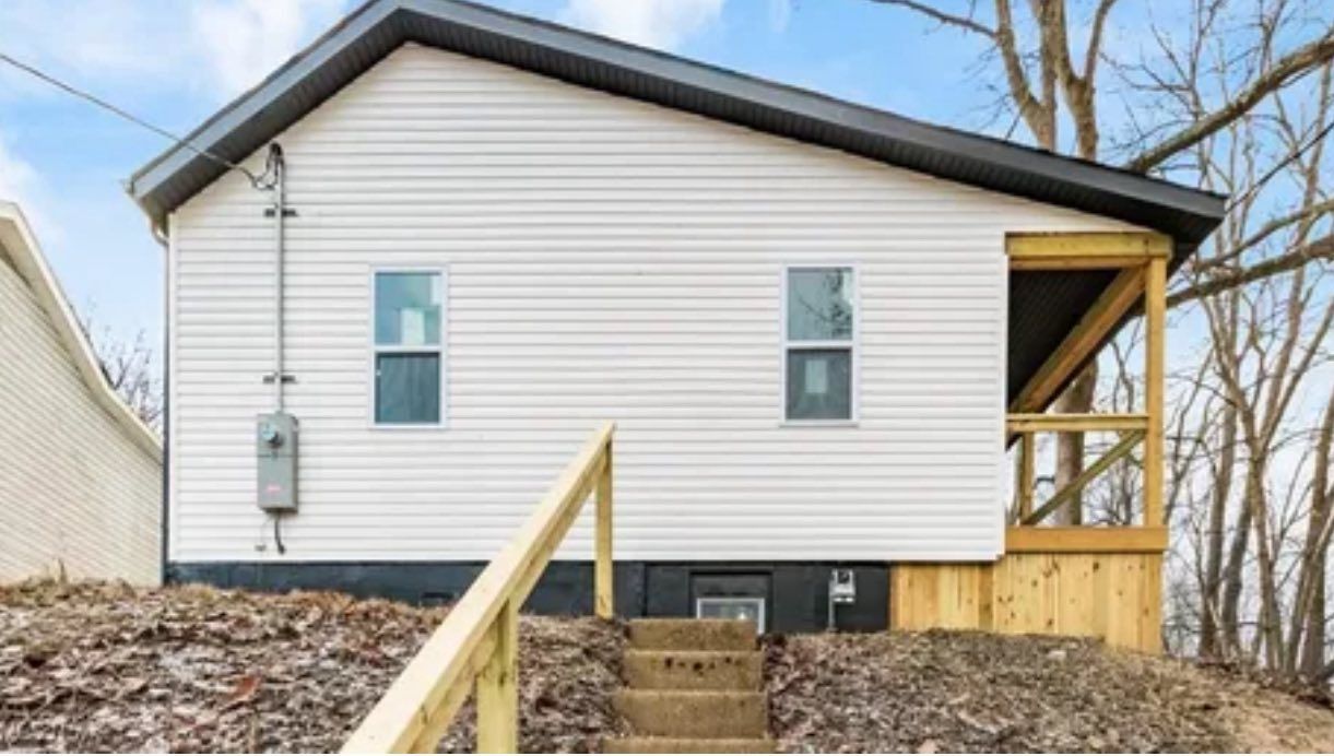 A white house with a dark grey roof and a wooden porch, featuring stairs leading up from a dirt slope with fallen leaves.