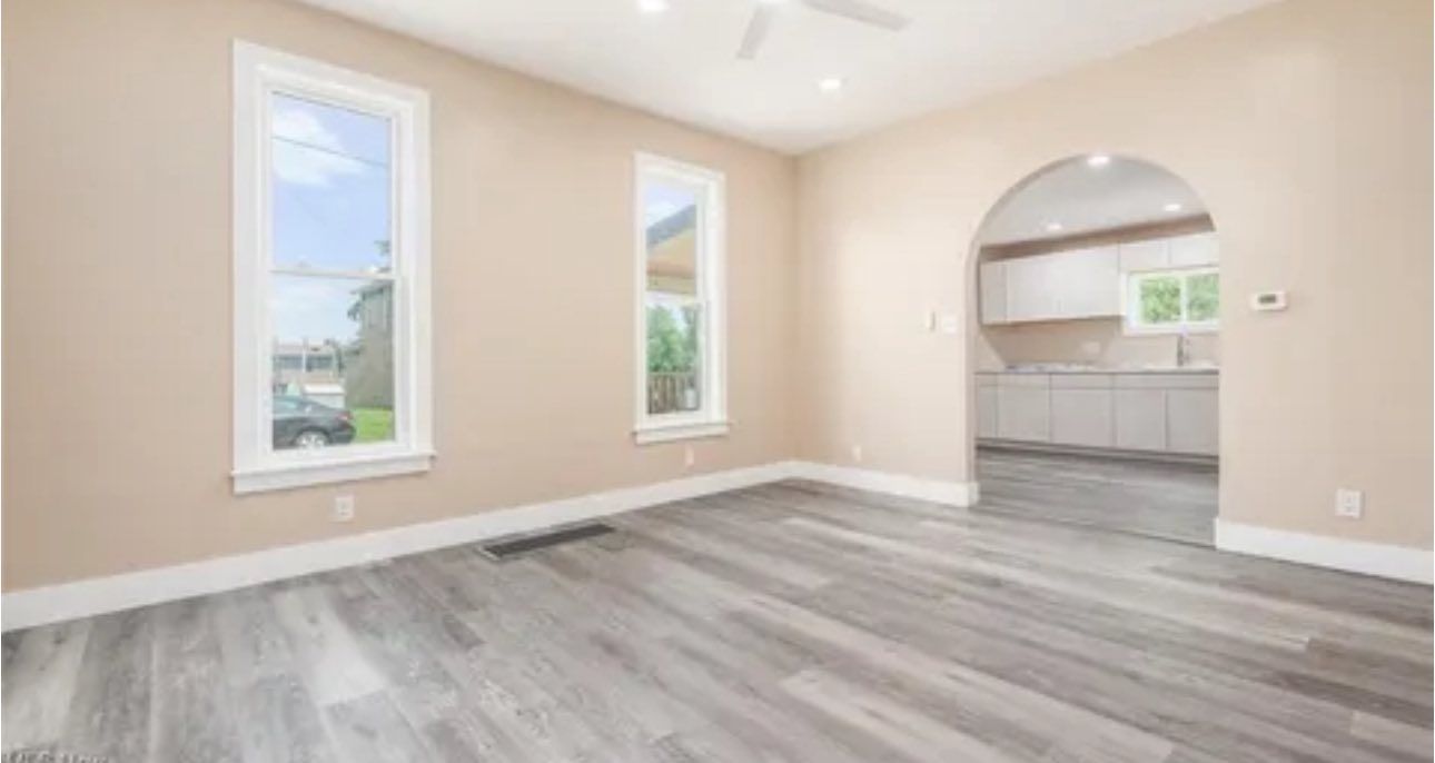 Bright, empty living room with light beige walls, wood-look flooring, two windows, and an arched entry to a kitchen.