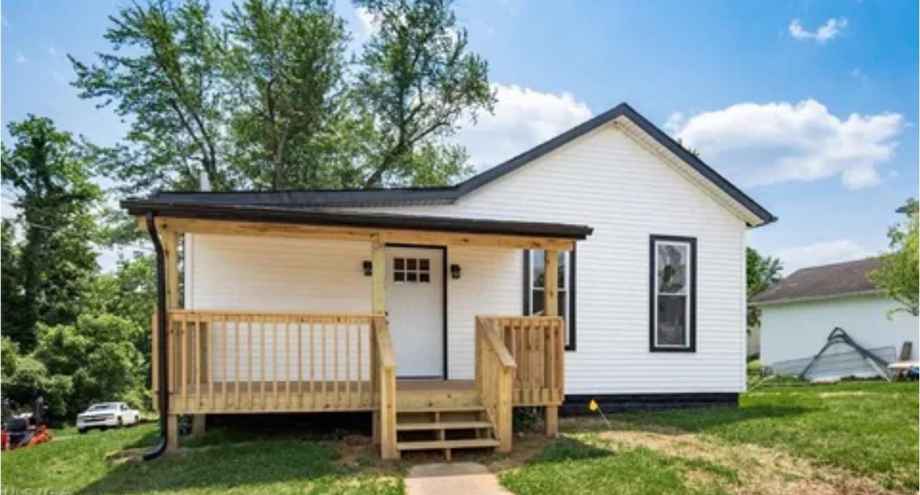 A small, white, single-story house with a black roof, dark window trim, and a newly built wooden front porch.