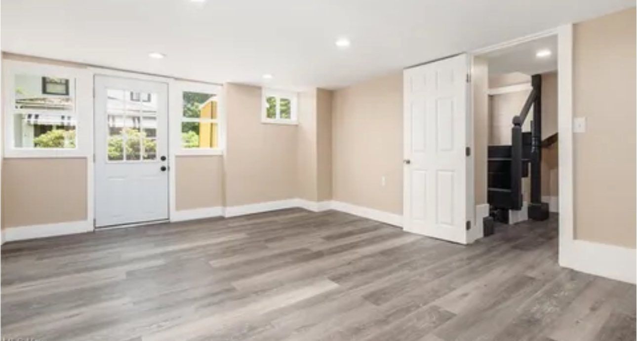 A bright, finished basement with light walls, wood-look flooring, a white exterior door, and an open doorway to stairs.